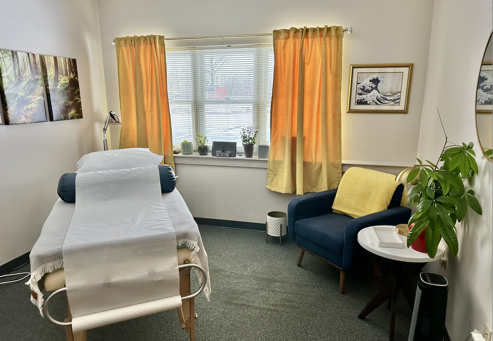 A room in a healthcare or massage setting with a single bed covered in white sheets, a black bolster pillow, a small side table, a blue armchair with a yellow throw blanket, a white round table with a plant and tray, and framed artwork on the walls. There are yellow curtains and a window with blinds, and indoor plants on the windowsill.