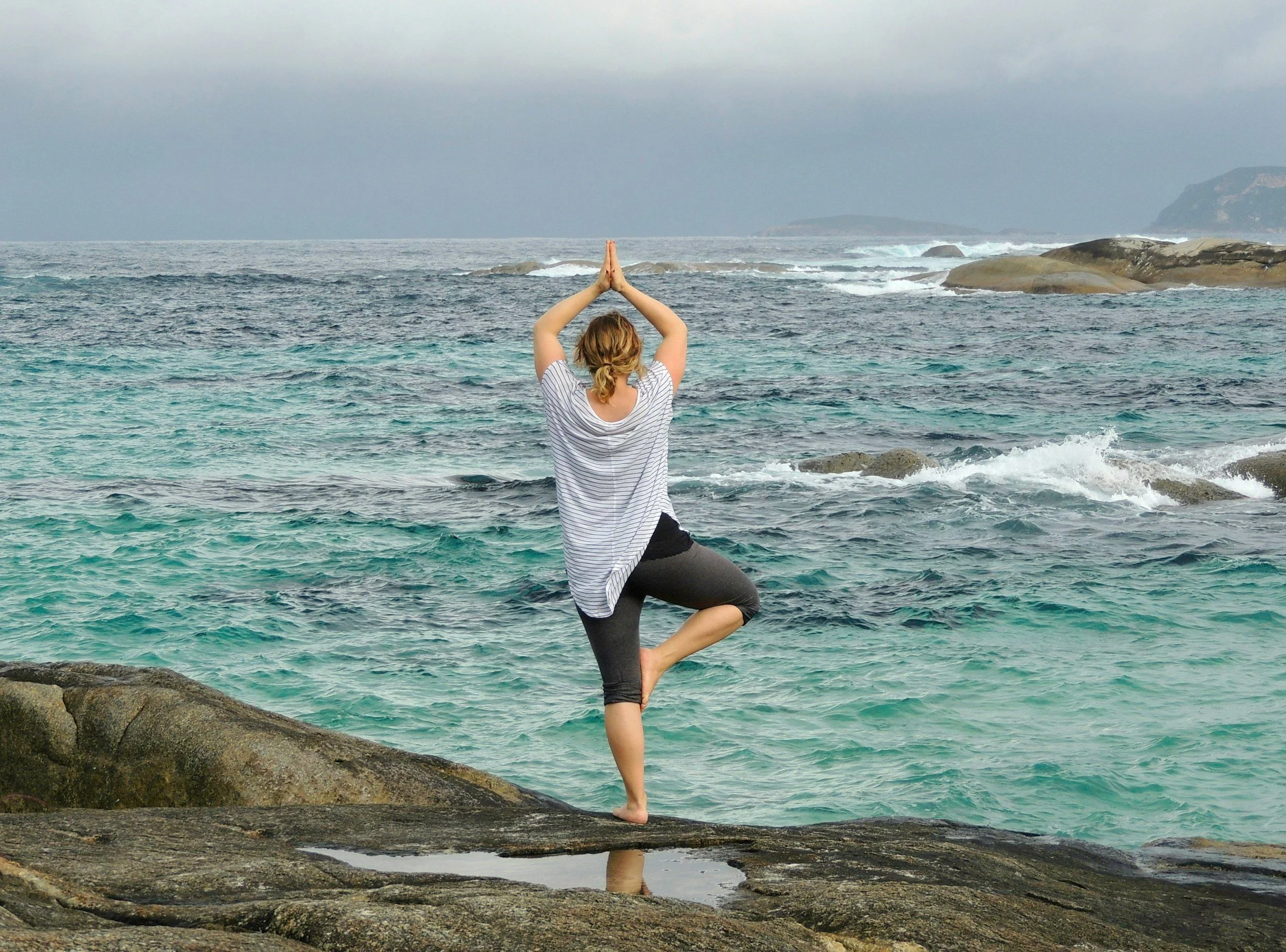 A woman practicing yoga on a rocky shoreline facing the ocean in a tree pose with hands together overhead.