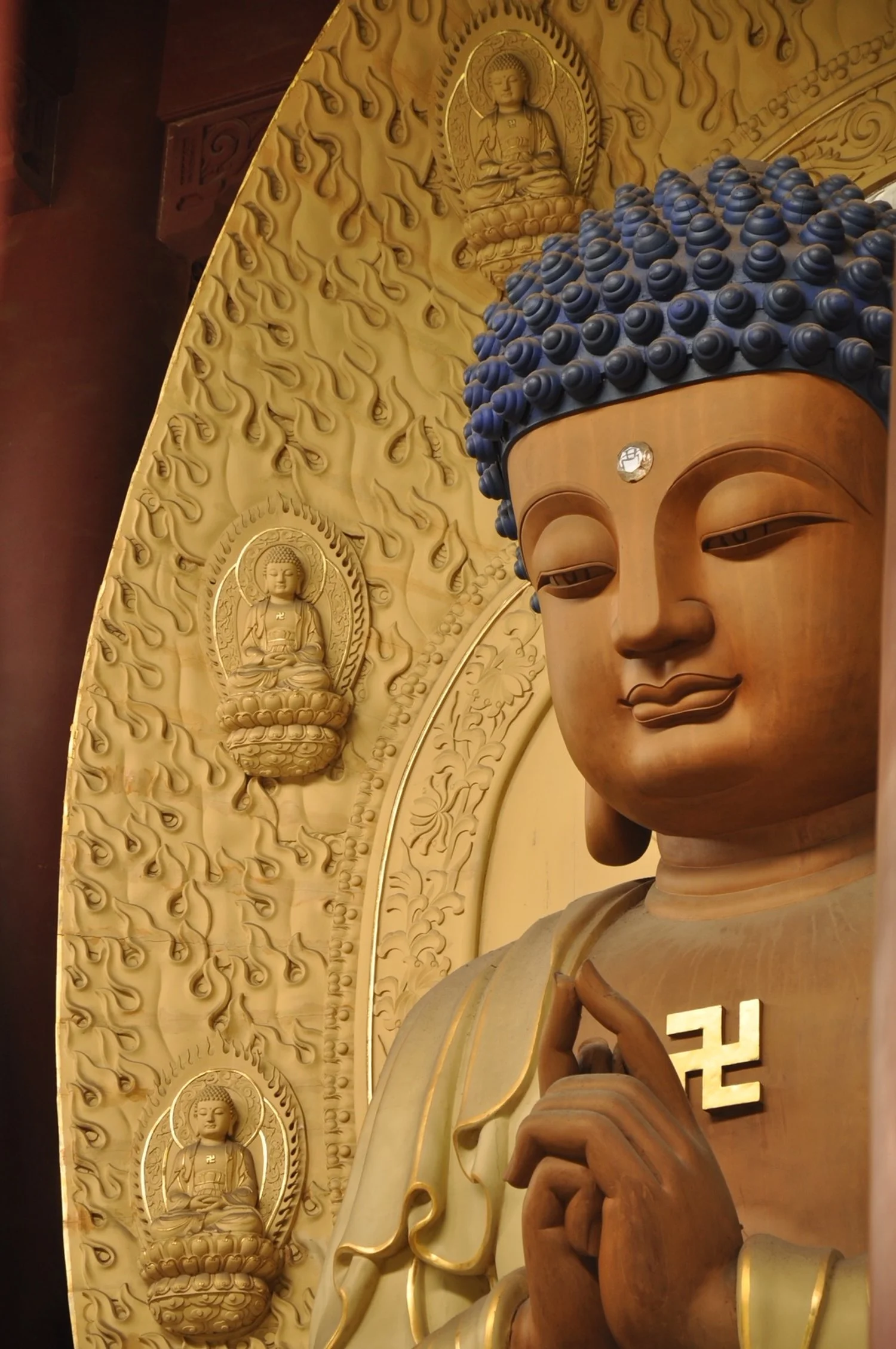 A large wooden statue of Buddha with blue hair and a serene expression, adorned with a small mirror and a swastika symbol on its chest, surrounded by ornate golden decorations and smaller Buddha figures engraved on the background.