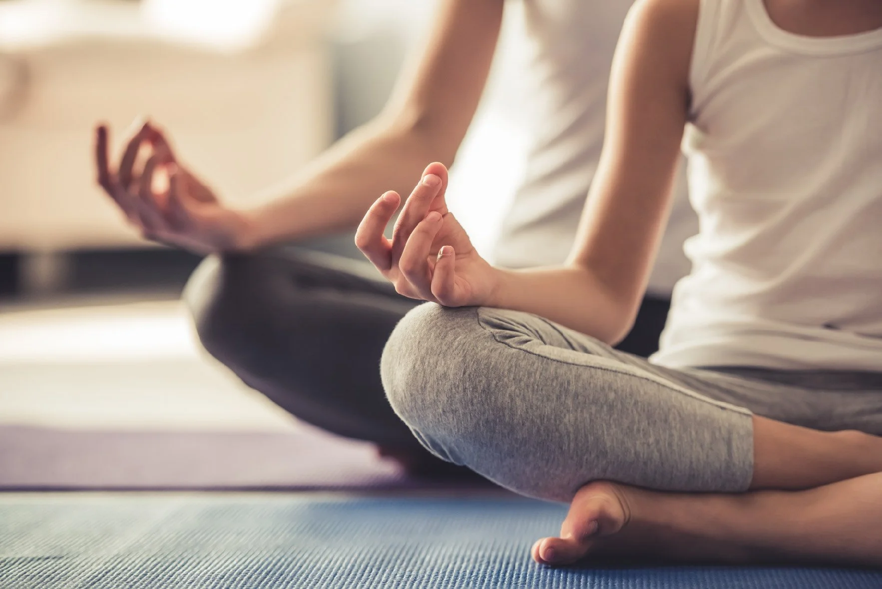 Close-up of two people practicing yoga or meditation, sitting cross-legged with one hand on their knee and fingers in a mudra, on yoga mats indoors.