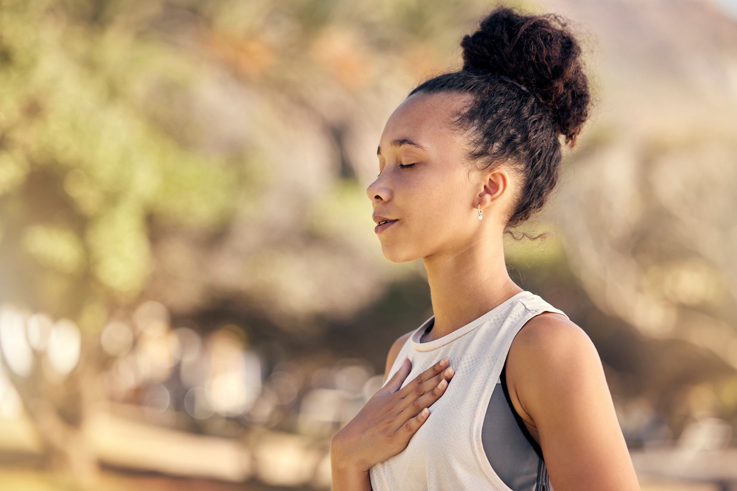 Young woman standing outdoors with her hand over her heart, eyes closed, wearing athletic clothing and a bun hairstyle, with a blurred natural background.