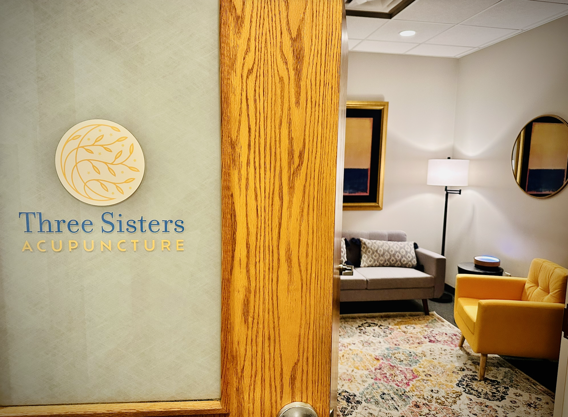 The door to Three Sisters Acupuncture with a view of a cozy waiting room with a gray sofa, yellow armchair, patterned rug, two framed pictures, a floor lamp, and an echo device.