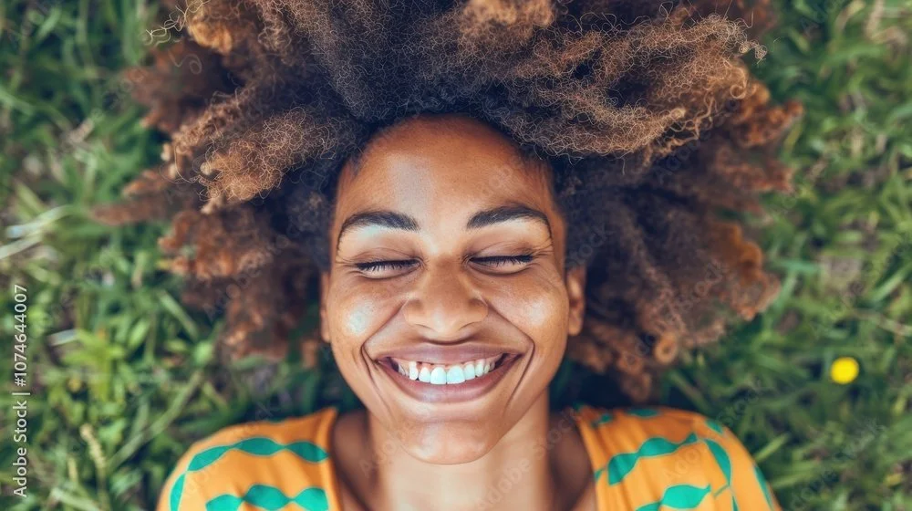 Young woman with curly hair lying on grass, smiling with eyes closed