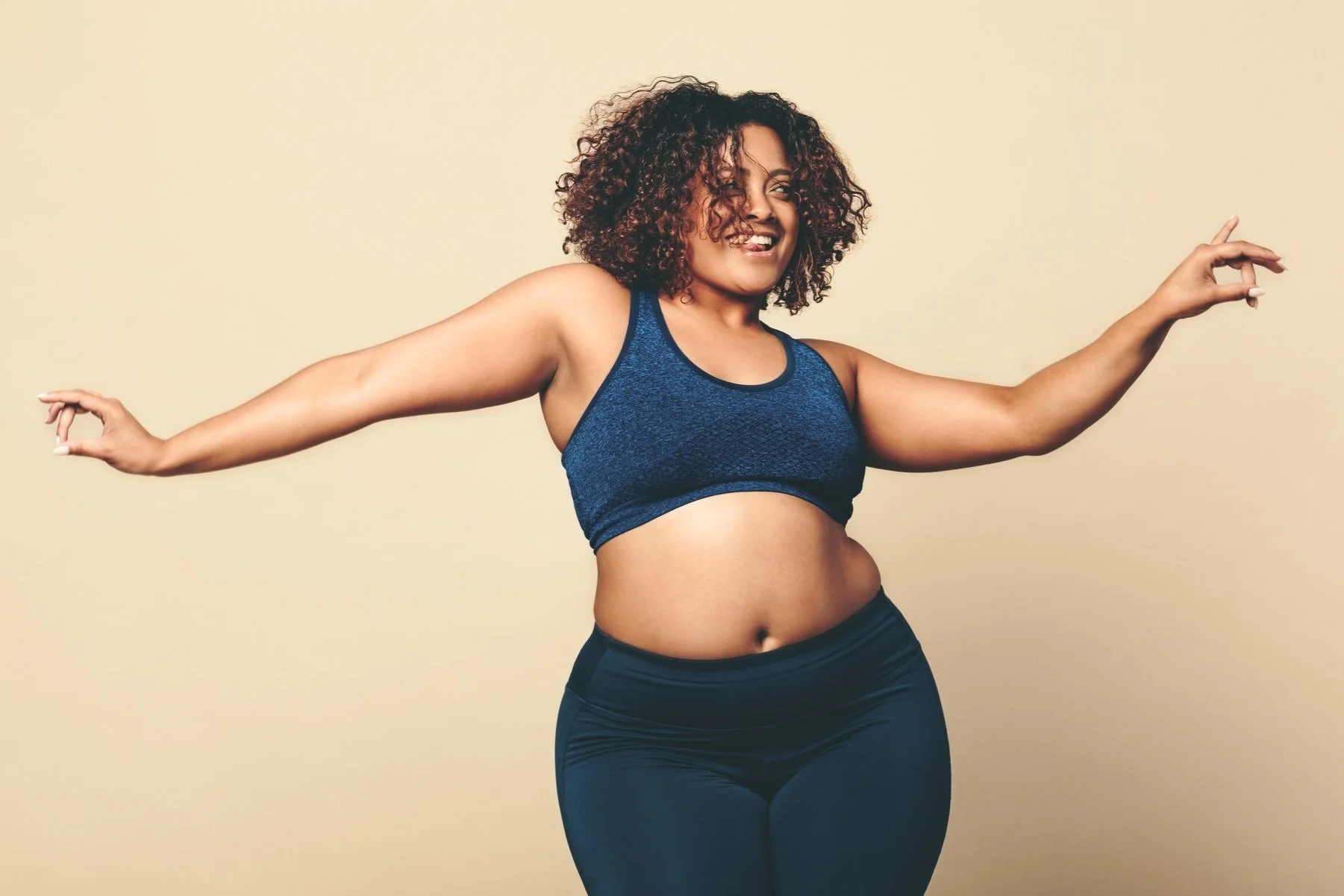 Smiling woman with curly hair wearing a blue sports bra and black leggings, arms extended to the sides, standing against a beige background.