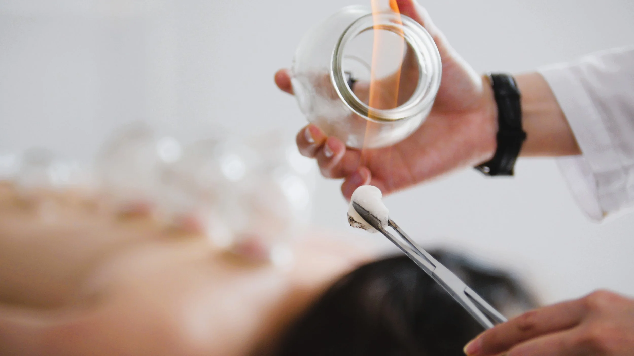 A person holding a small jar and a cotton swab, with another person's head in the foreground.