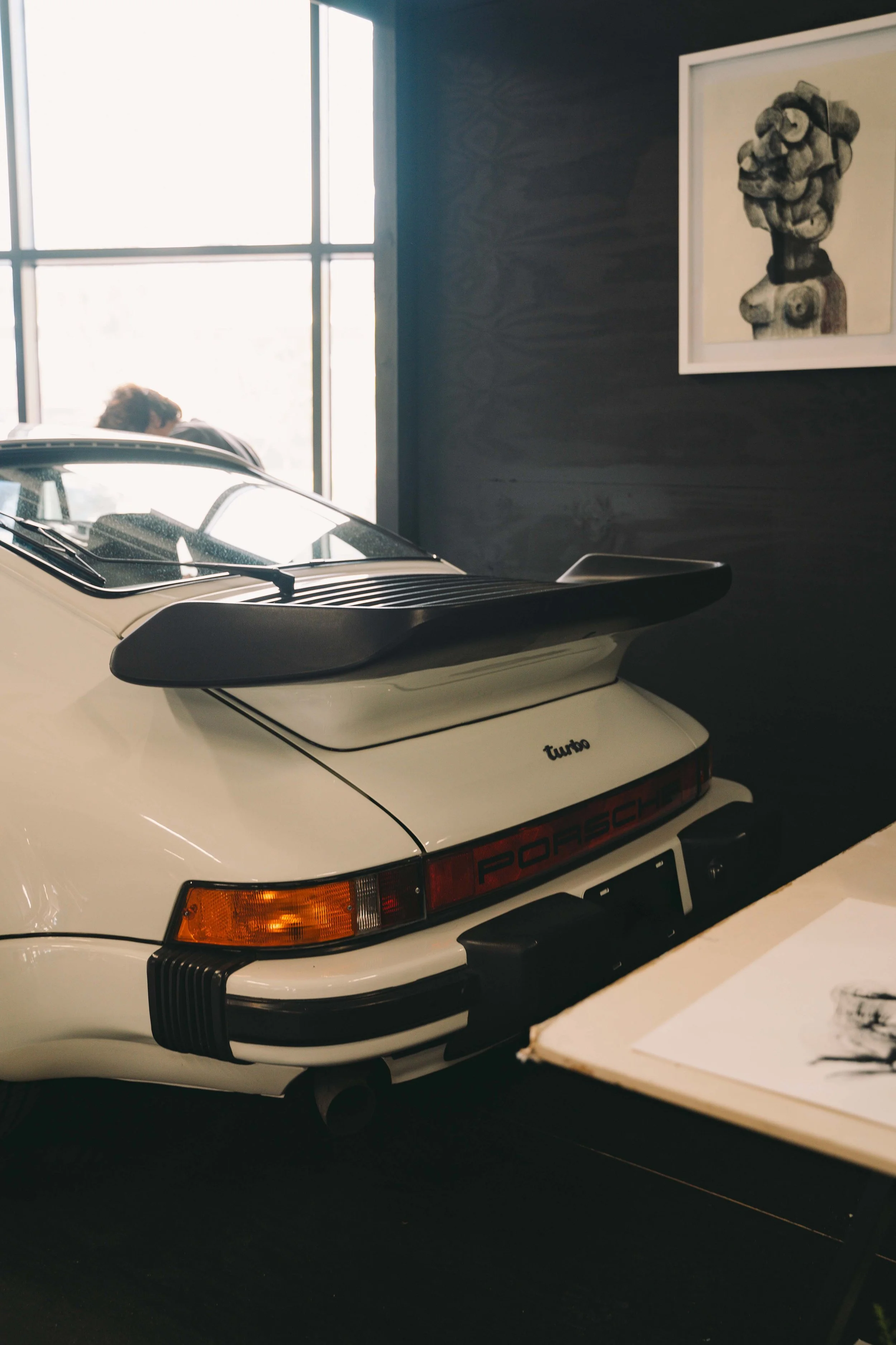 Rear view of a white Porsche 911 Turbo with a black rear wing, parked indoors near a black wall with a framed artwork, and a person near a window in the background.
