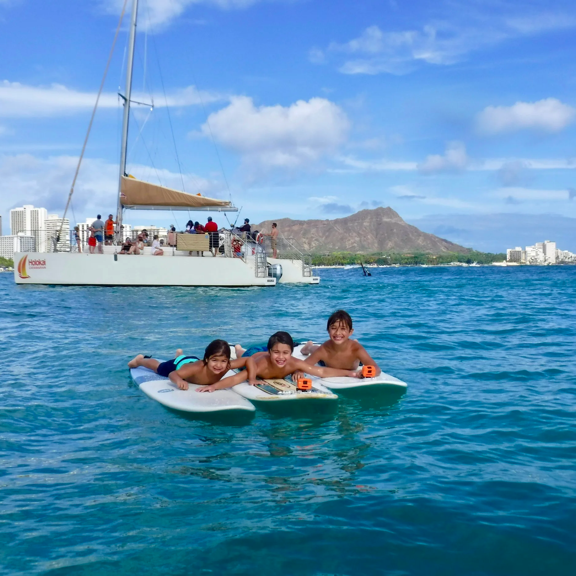 Three brothers, Hook-Up-Surfing beginner surf students are lying on surfboards in the water, smiling, with a sailboat, the Waikiki skyline with Daimondhead in the background under a partly cloudy Waikiki sky.