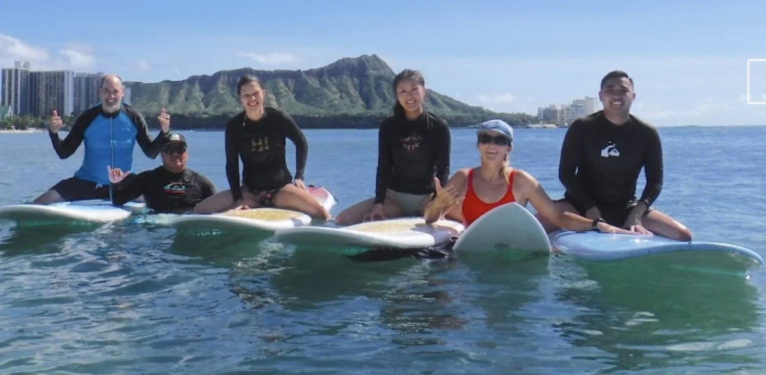 Family out in Hawaii for a surf lesson with Hook Up Surfing, sitting on surfboards on a clear sunny day in Waikiki with Diamondhead in the background.