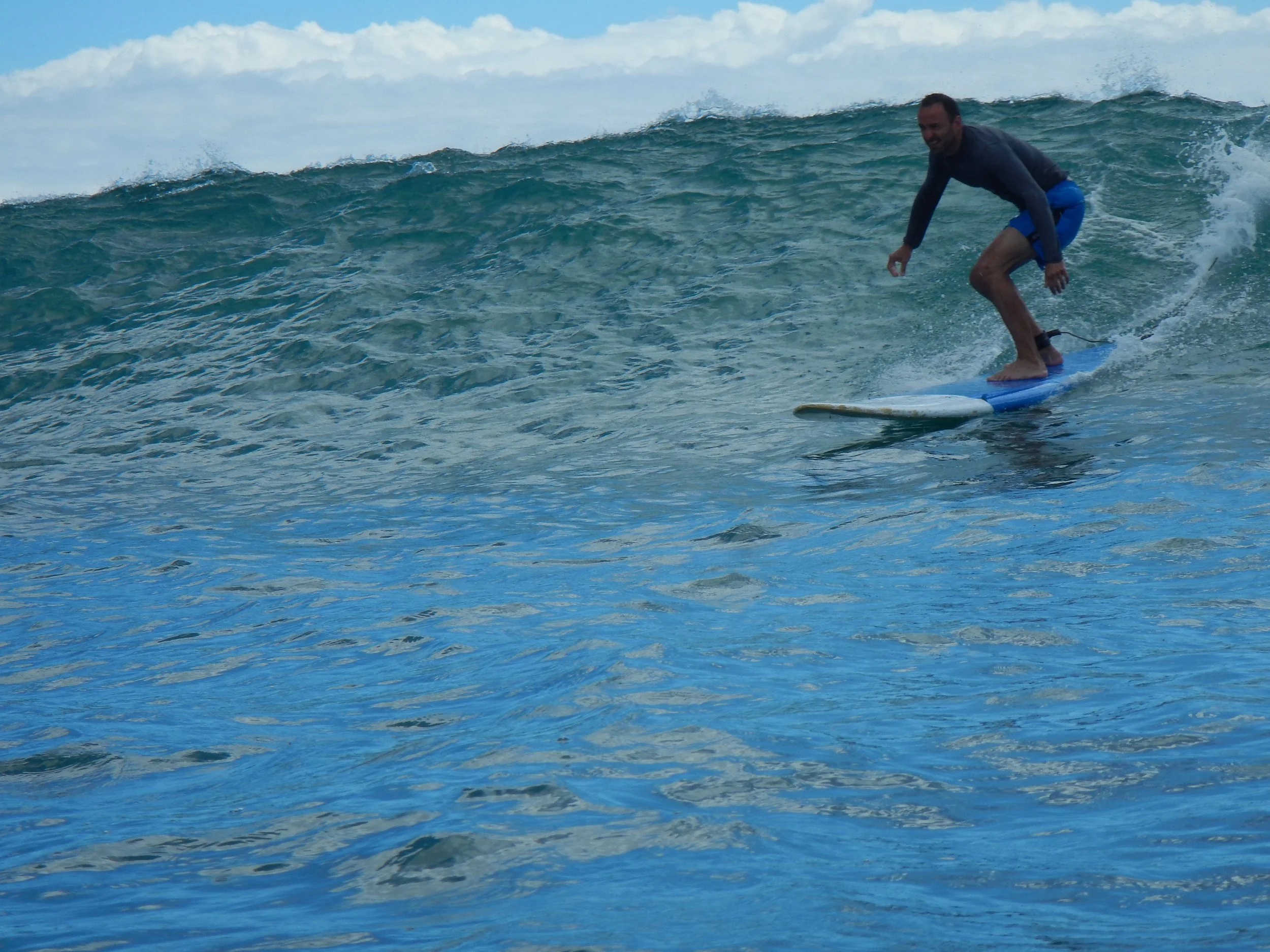 A Hook Up Surfing surf student surfing on a wave in the Waikiki, wearing a black long-sleeve shirt and blue shorts, smiling and balancing on his surfboard.