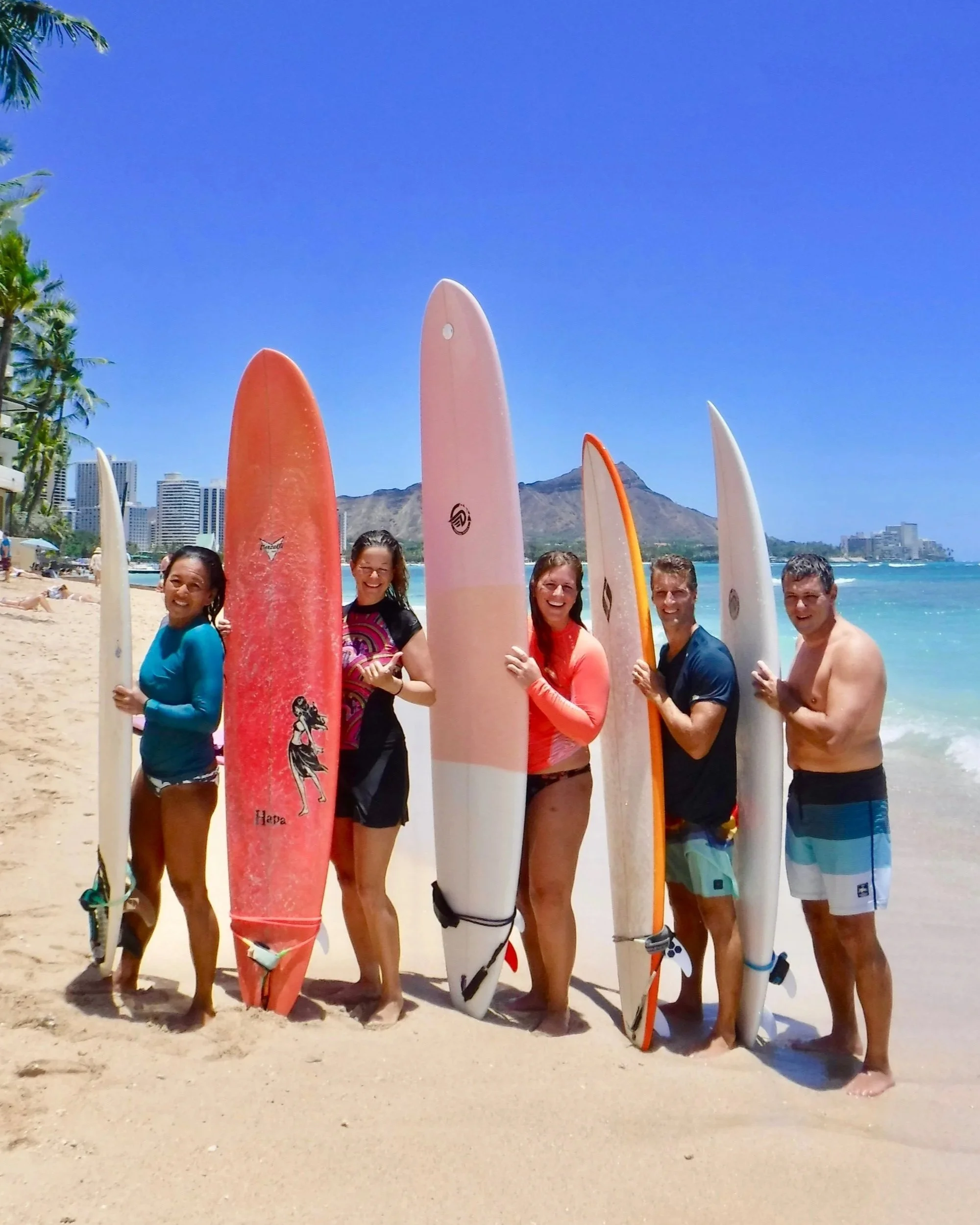 Five real surf coaching students posing together on the beach after a session in Waikiki beach holding surfboards, smiling.