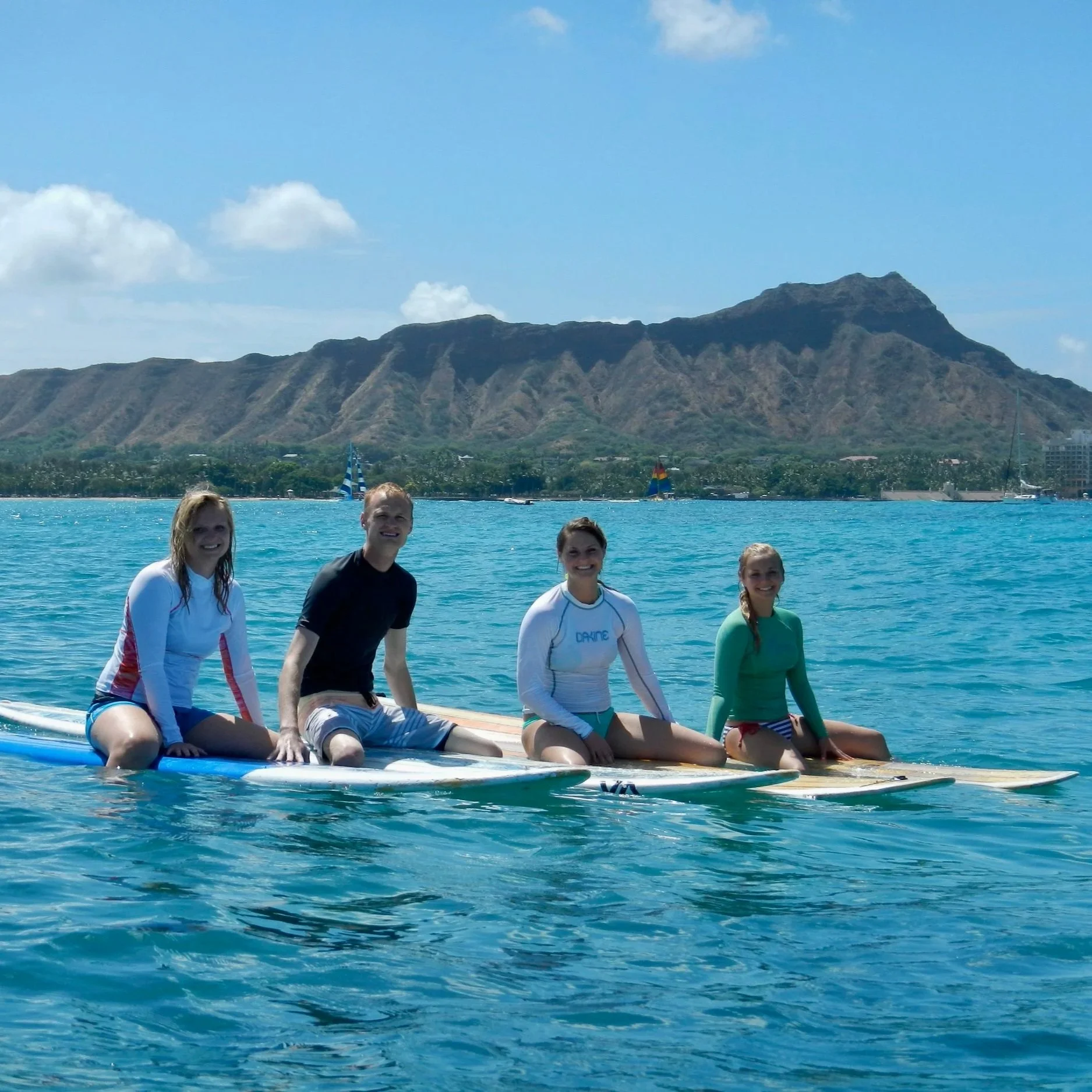 Family of four people, Hook-Up-Surfing beginner surf students, are sitting on surfboards in the water, smiling, with a sailboat, the Waikiki skyline with Daimondhead in the background under a partly cloudy Waikiki sky.