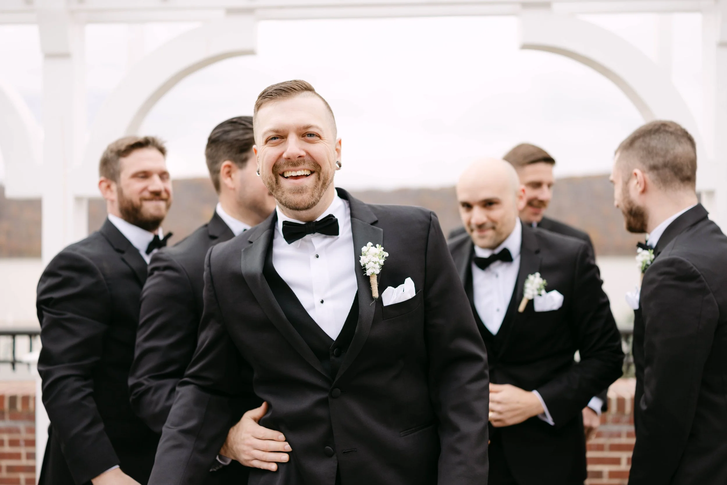 Group of men in tuxedos at outdoor wedding, with one smiling in front, with a backdrop of a white structure and overcast sky.