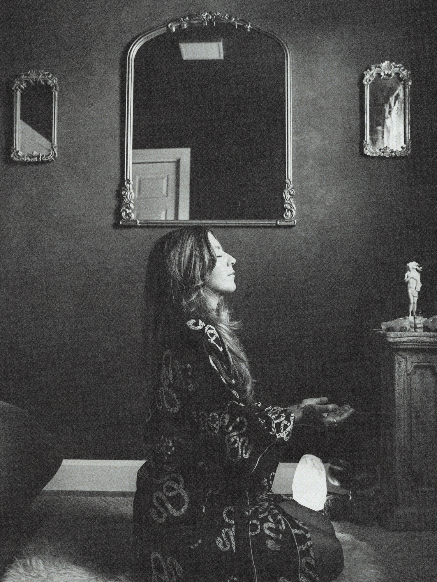 A woman with long hair sitting cross-legged on the floor in meditation, with eyes closed, in a room with dark walls. Behind her is a large ornate mirror, and two smaller mirrors hang on either side. A statue on a table is on the right side of the image, with a glowing light on the floor near her.