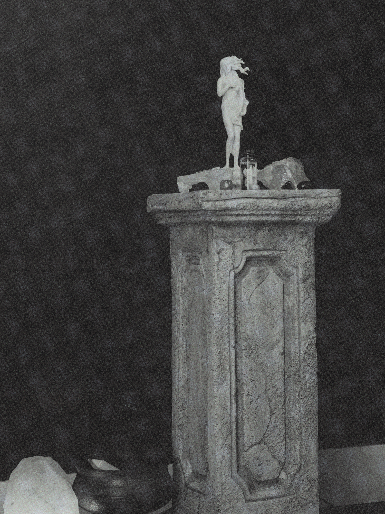 A stone pedestal with a small sculpture of a standing woman and a glass bottle on top, against a dark background.