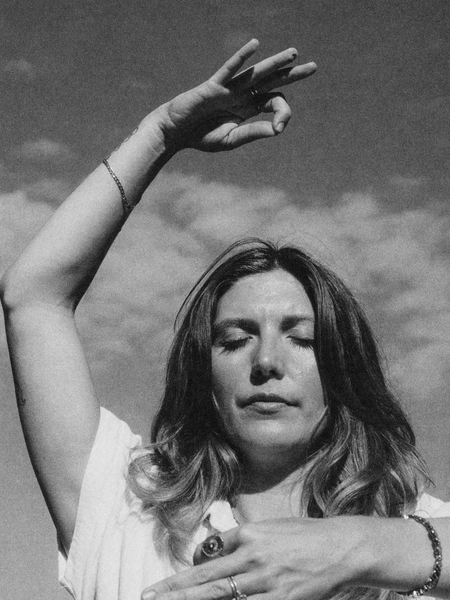 A woman with shoulder-length hair and closed eyes, raising one arm with her fingers forming a gesture, outdoors against a cloudy sky.