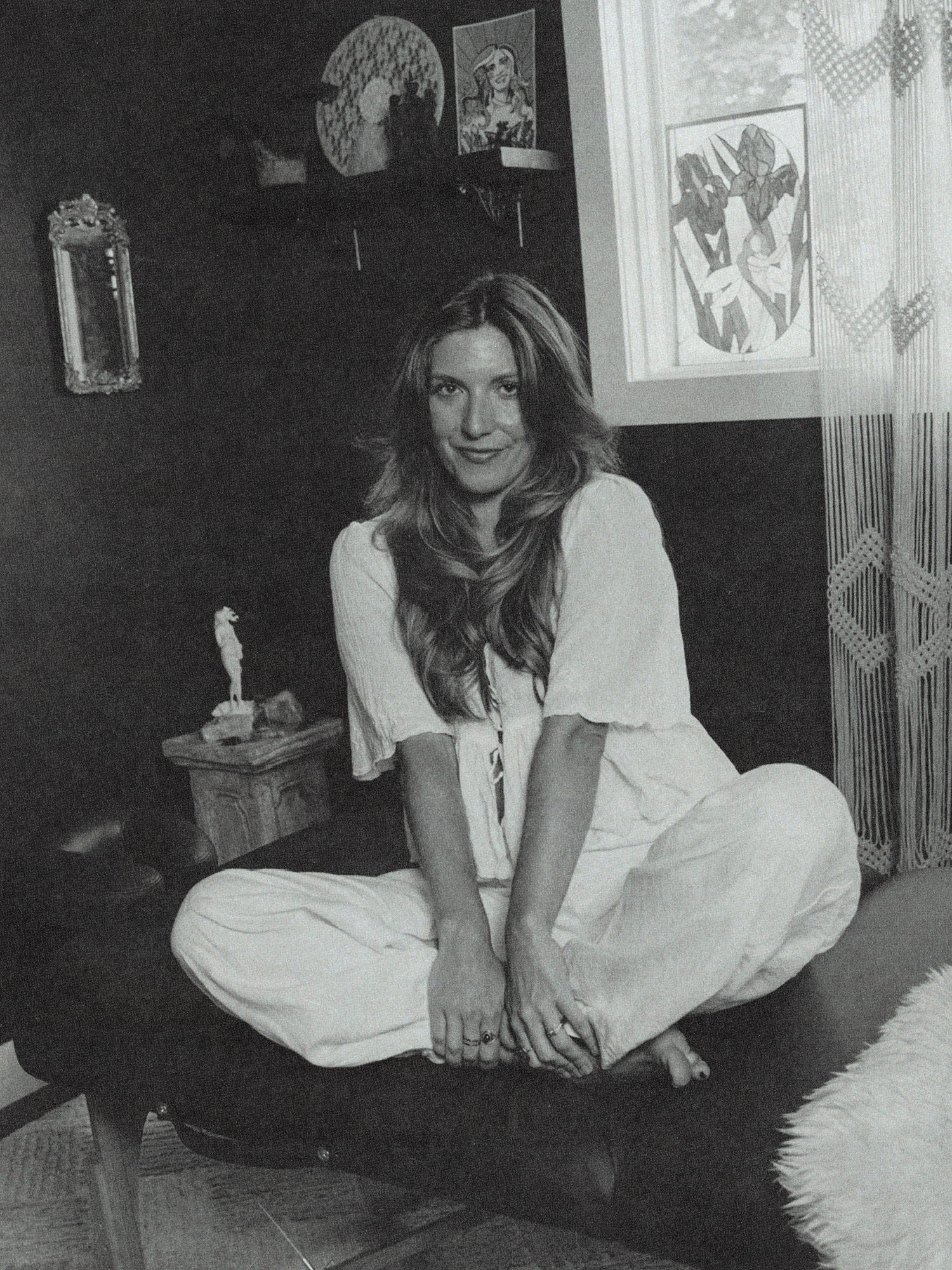 Woman with long hair sitting cross-legged on a chair in a room with dark walls, surrounded by artwork and a window with patterned curtains.