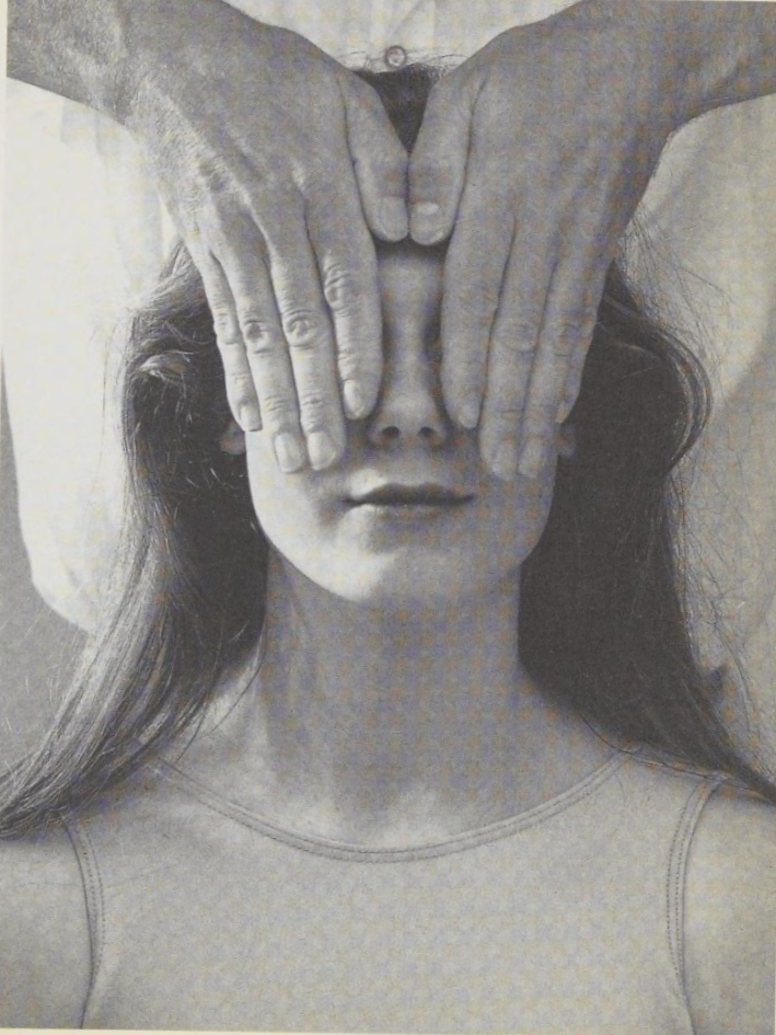 A black-and-white photograph of a woman with long hair, wearing a sleeveless top, with a pair of adult hands covering her eyes.