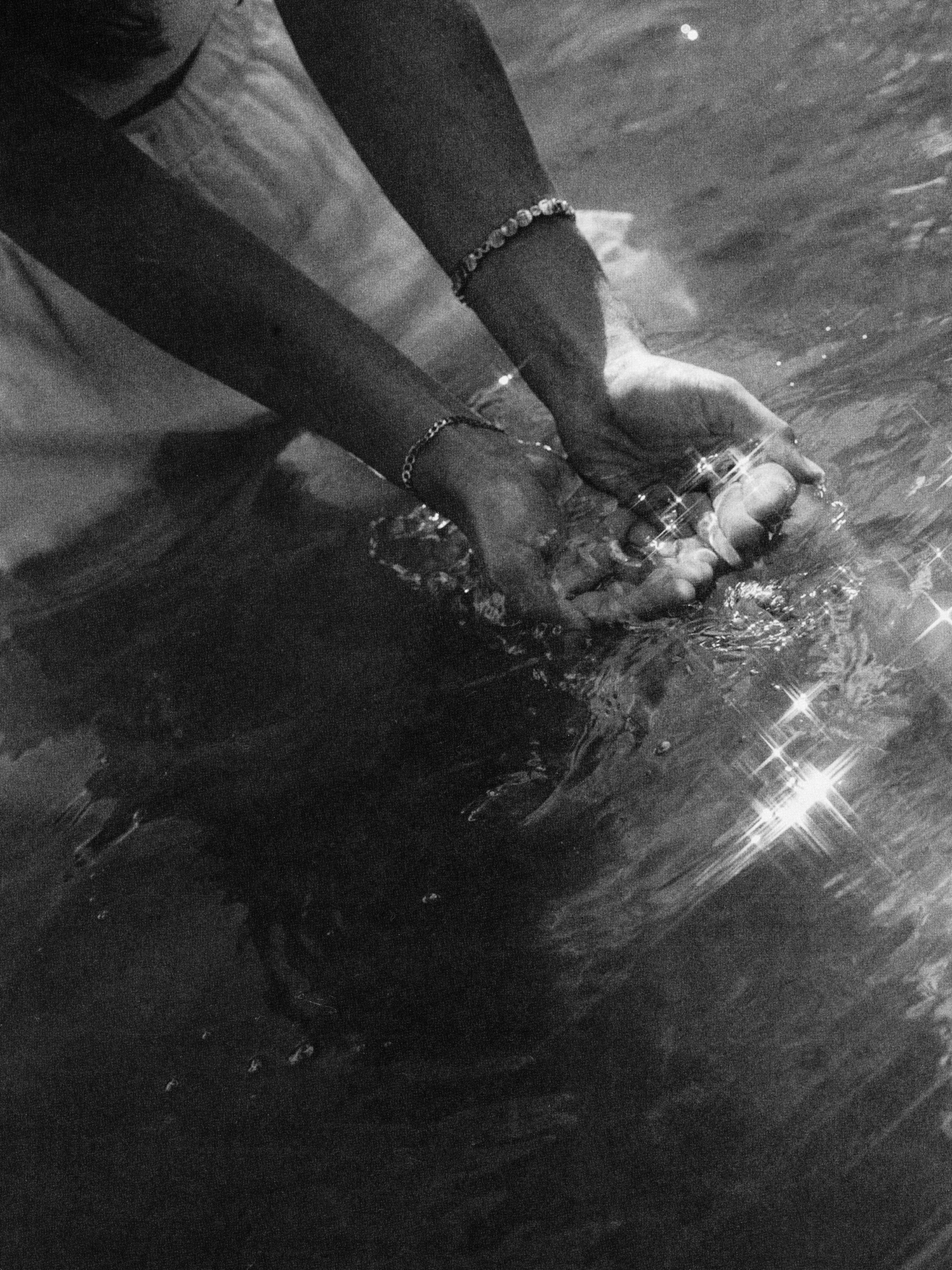 Hands held together in water, wearing bracelets, with sunlight reflecting on water's surface.
