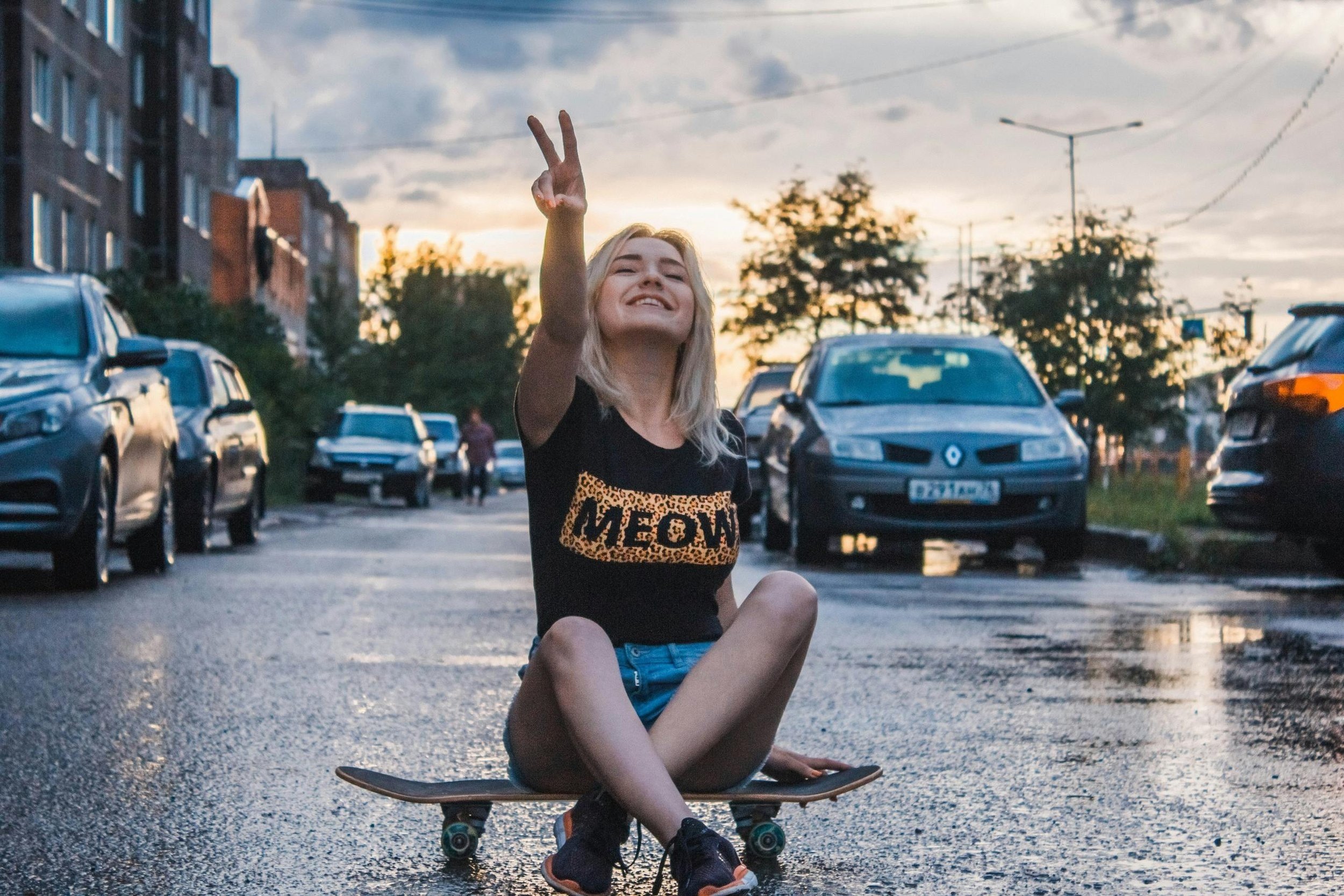Mujer joven sonriendo, sentada en una patineta en la calle, haciendo el signo de la paz con los dedos con coches estacionados y un cielo nublado al fondo.