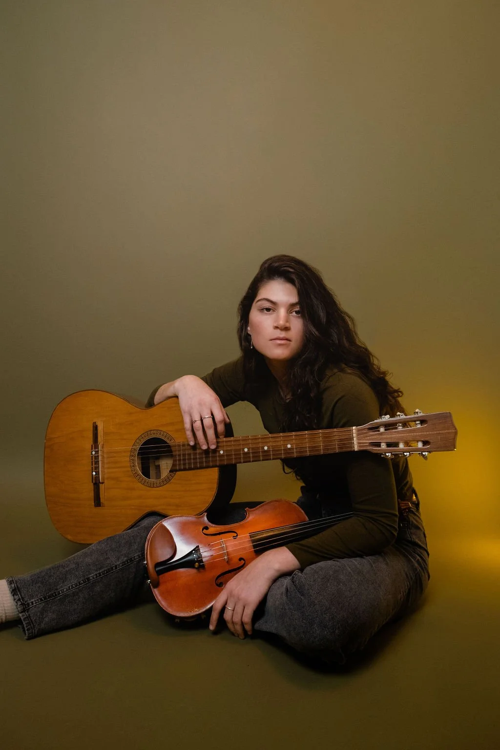 A young woman with long dark curly hair, sitting on the floor against an olive green background, holding an acoustic guitar and a violin.