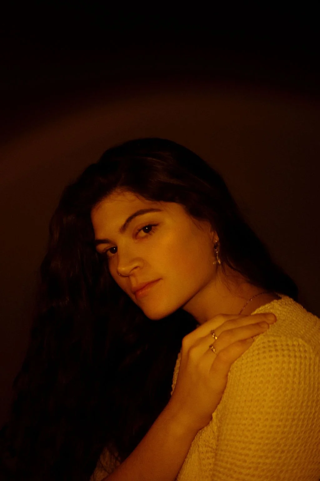 A woman with long dark hair gazing at the camera, wearing a yellow sweater, jewelry including a ring and earrings, against a dark background.