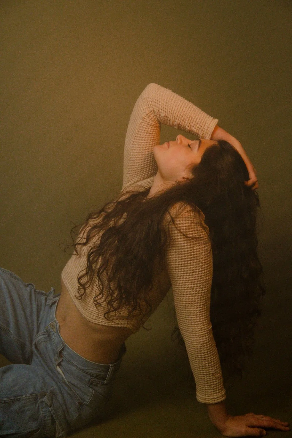 A woman with long, curly dark hair, wearing a beige checkered long-sleeve top and light blue jeans, is sitting on the floor against a plain olive-green background. She is posing with her head tilted back, eyes closed, one arm resting behind her head,