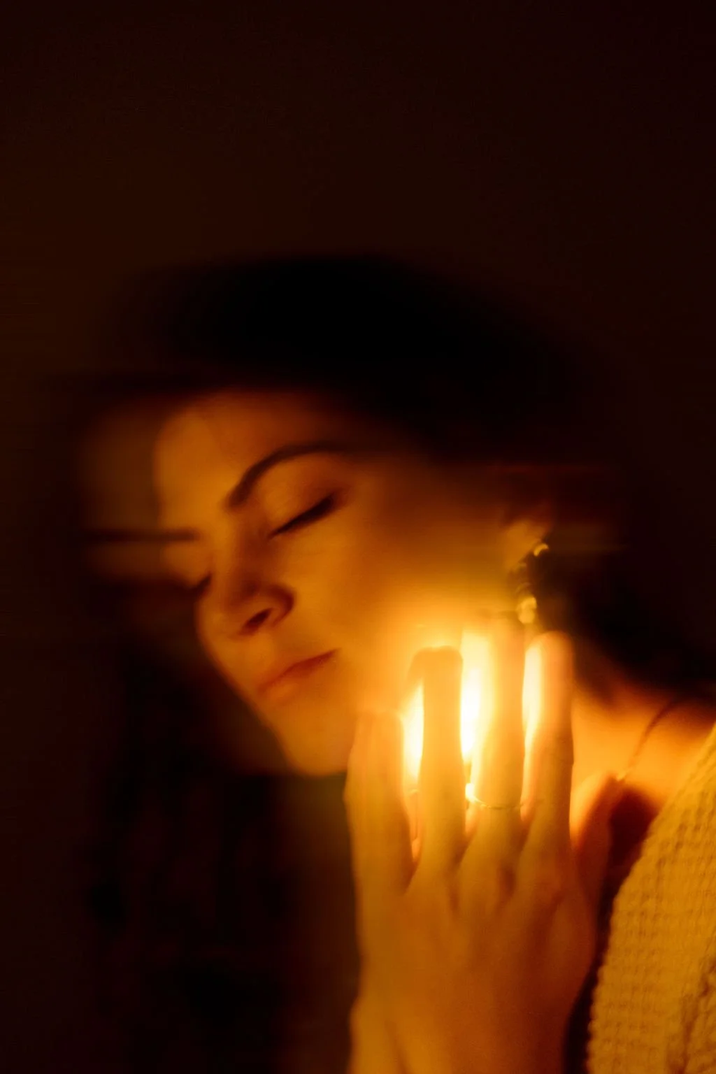 A woman with closed eyes holding a glowing candle close to her face in a dark environment.