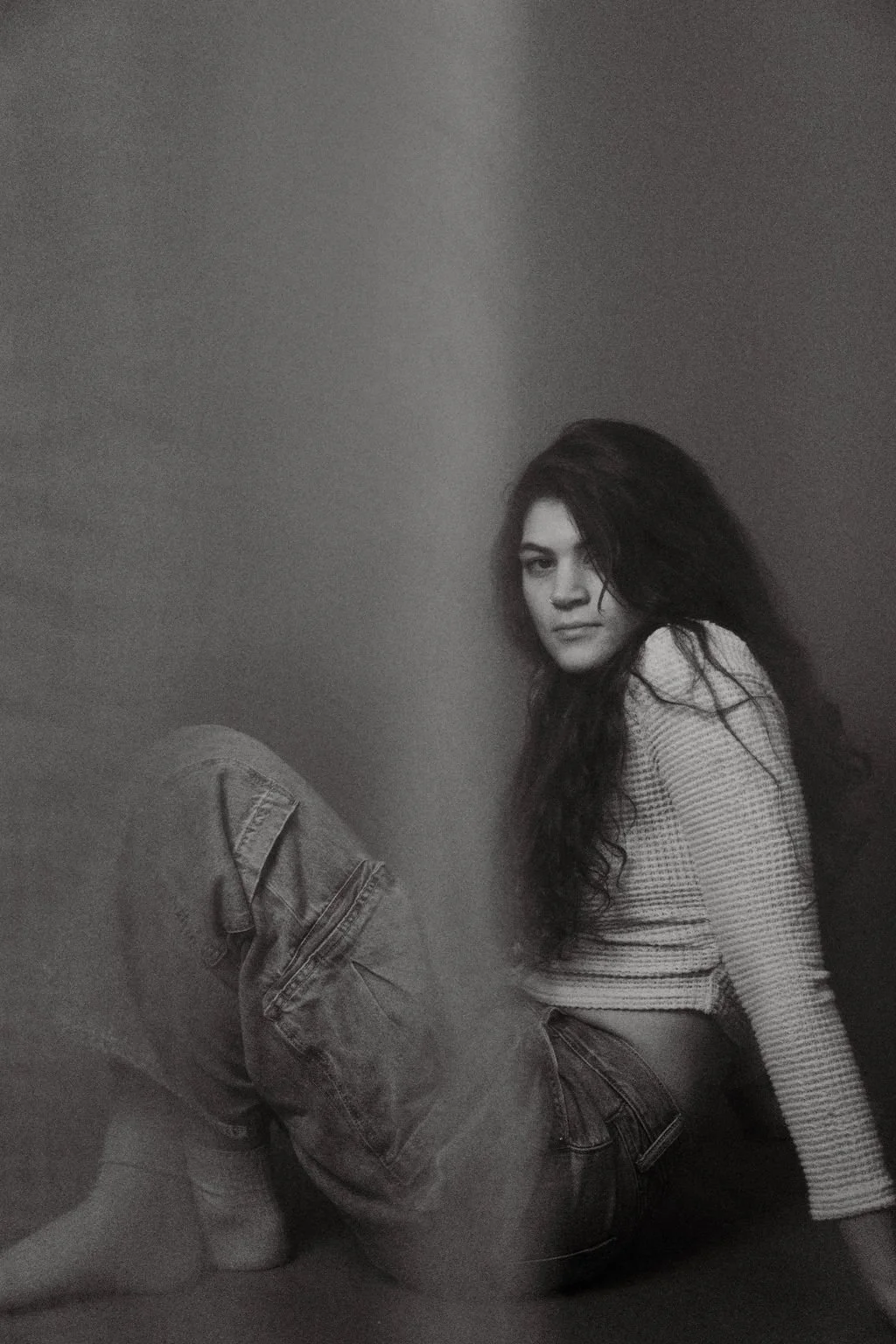 A young woman with long dark hair sitting on the floor in front of a plain wall, looking at the camera with a serious expression, wearing a striped long-sleeve top and jeans.