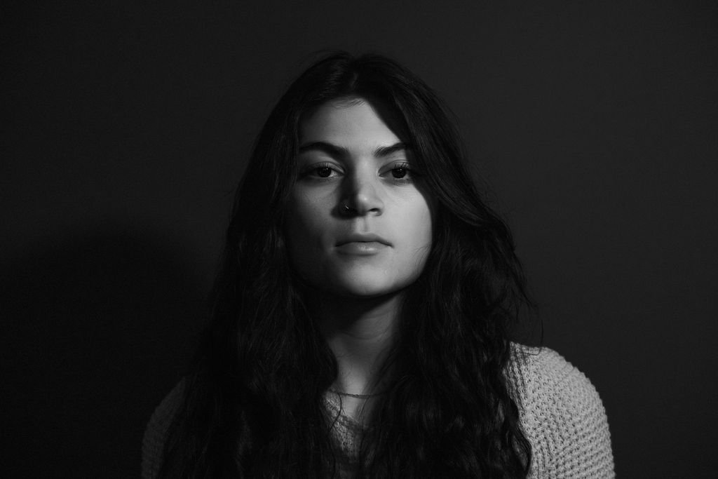 Black and white portrait of a young woman with long, dark, wavy hair, wearing a light-colored knitted sweater, against a dark background.