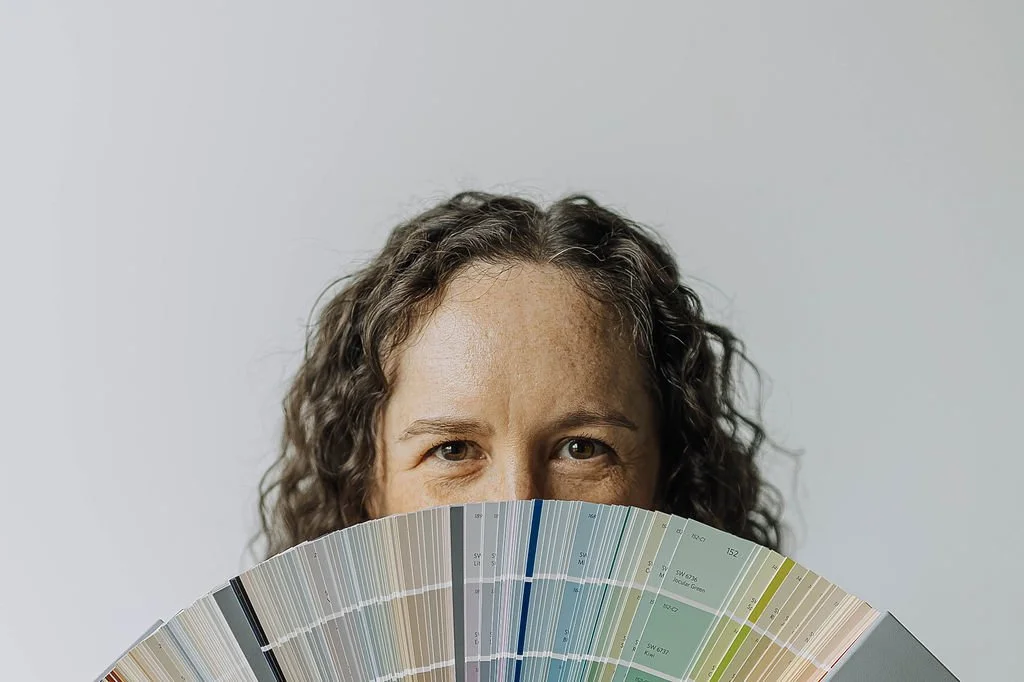 Anna Kuttel holding a color swatch fan in front of her face, revealing her eyes and forehead, with a plain light gray background.