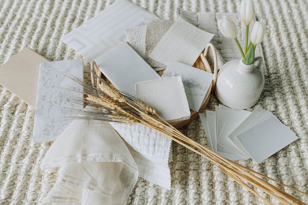 A collection of neutral-colored home decor items, including fabric swatches, a white vase with tulips, and woven textiles, arranged on a textured cream rug.