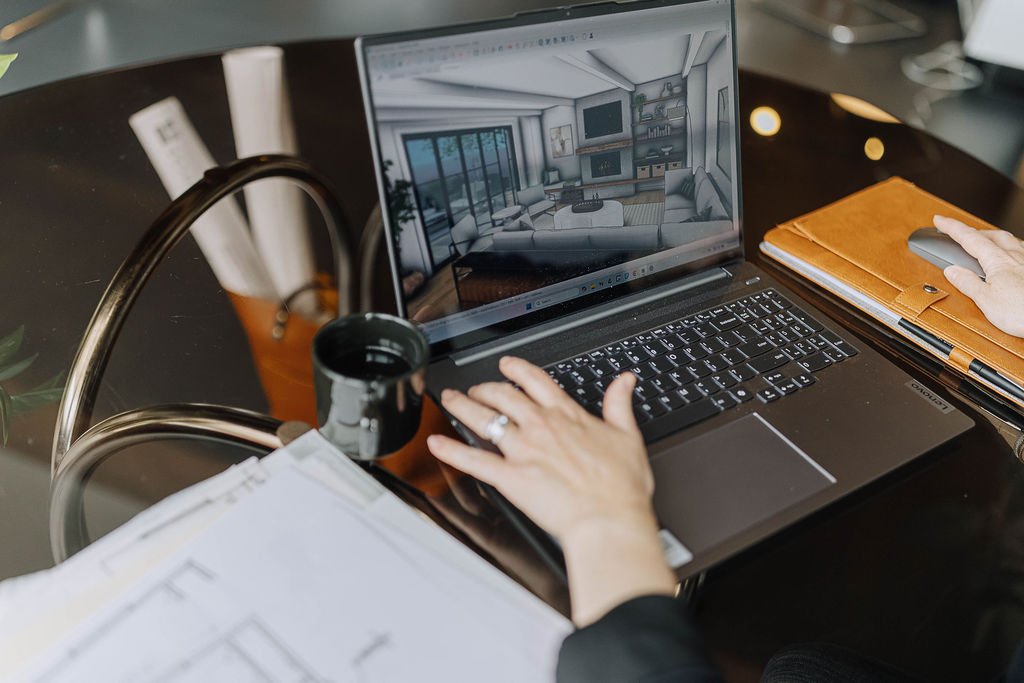 Anna Kuttel working on a laptop at a desk, viewing a 3D interior design of a living room on the screen. The desk has a cup, an orange notebook, a computer mouse, and some rolled-up papers.