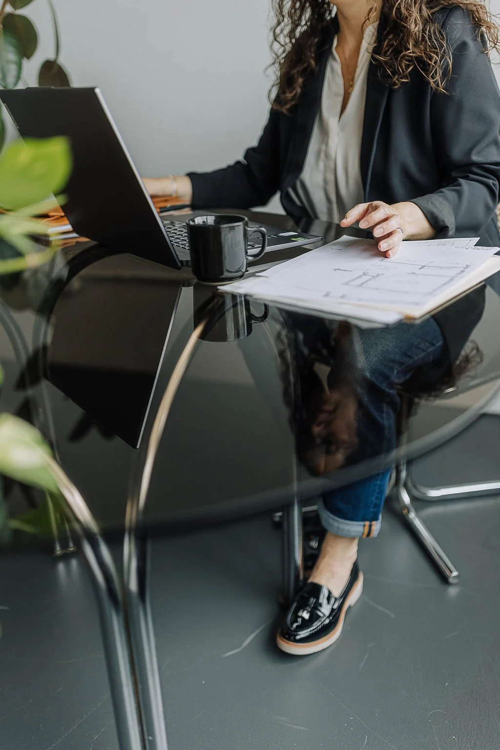 Anna Kuttel sits at a black glass-topped desk with a laptop, a black coffee mug, and architectural plans. She wears a blazer, white blouse, jeans, and loafers, with a plant partially visible in the foreground.