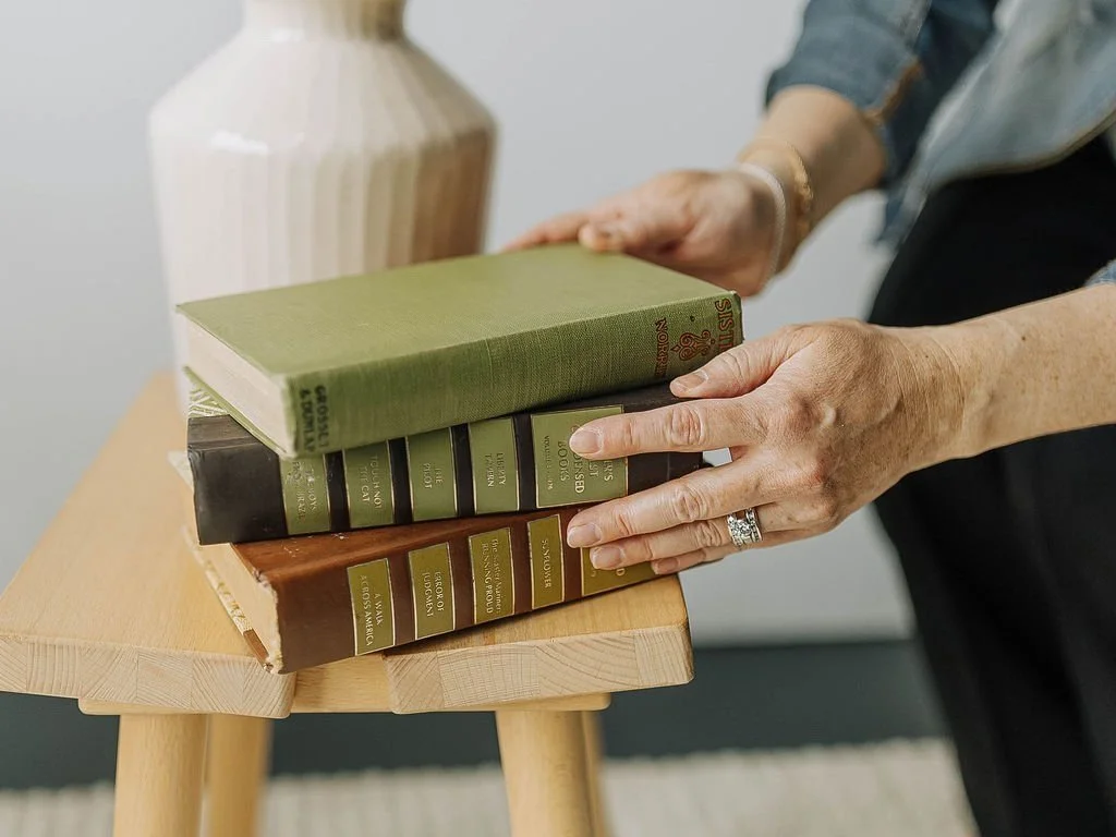 Anna Kuttel placing a green hardcover book on top of a stack of four books on a small wooden table, with a ceramic vase in the background.