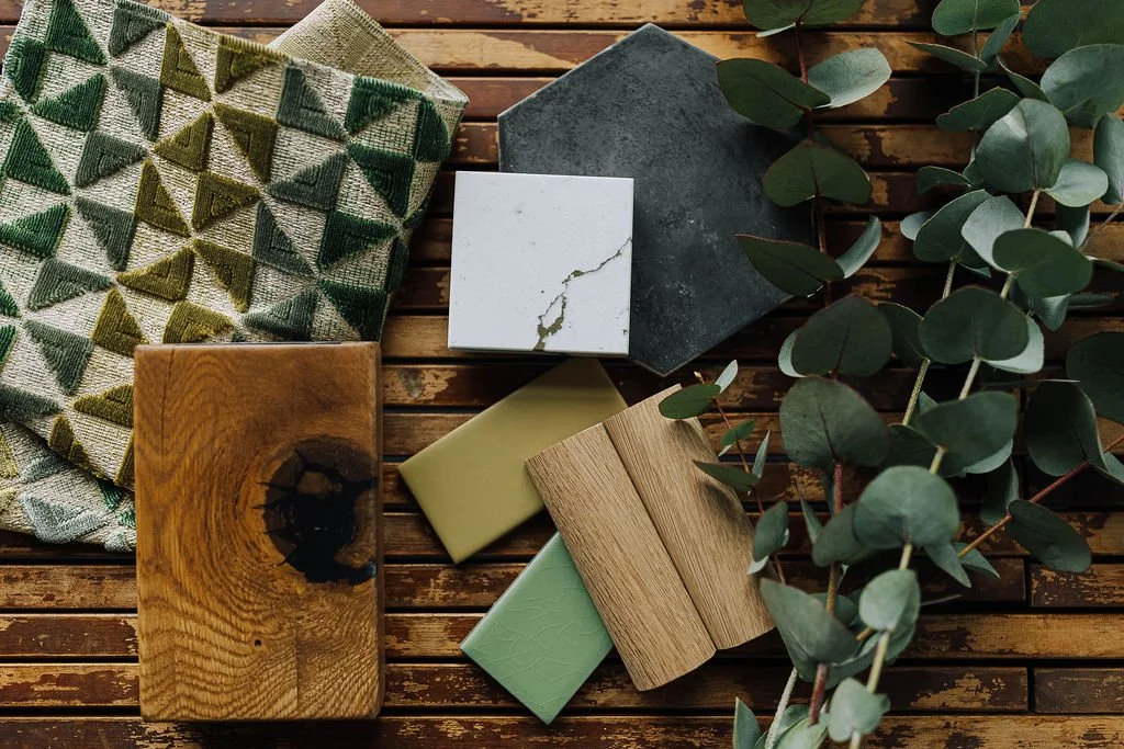 Collection of fabric swatches, tiles, wood pieces, and eucalyptus leaves on a wooden surface.