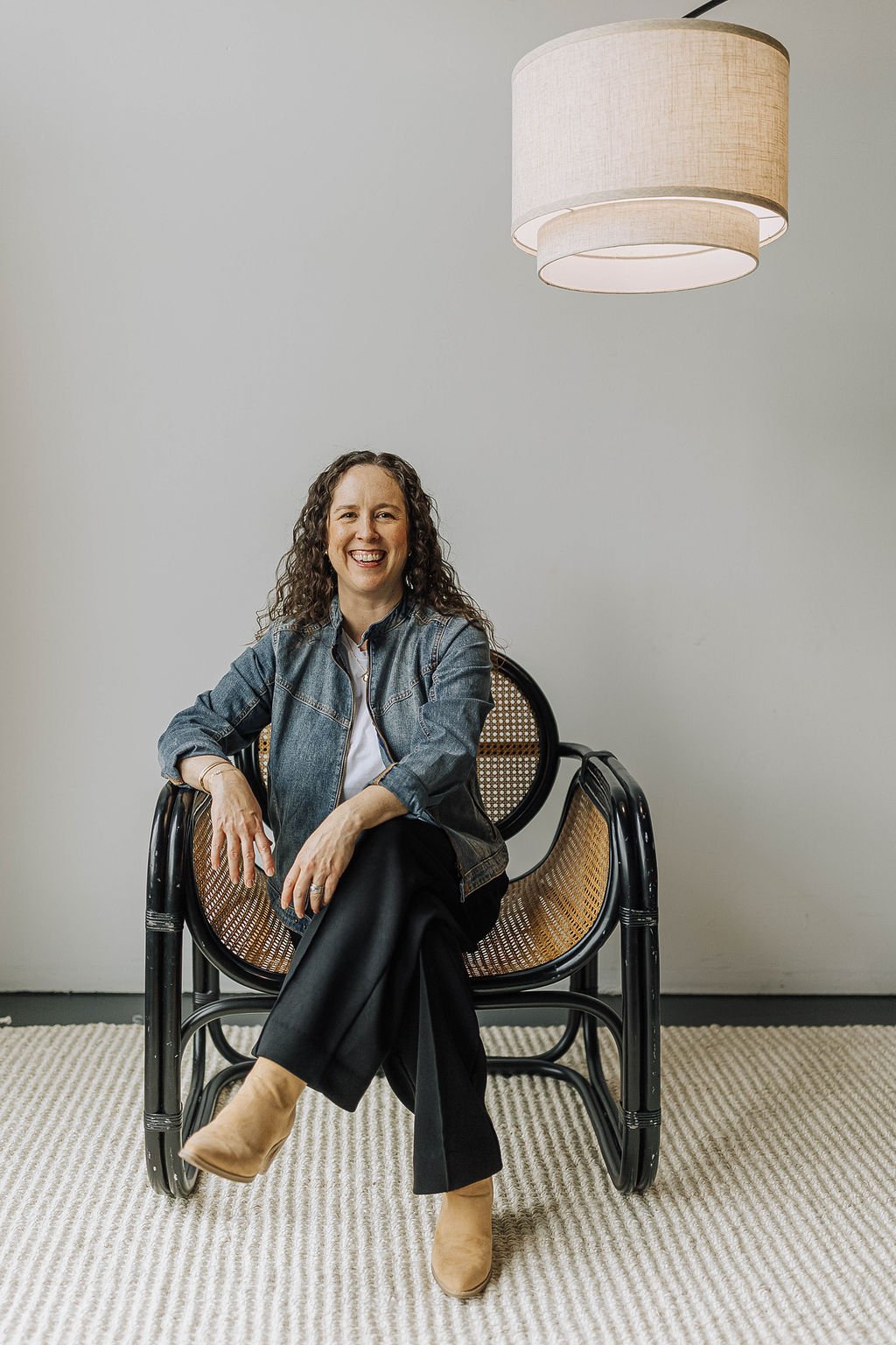 Anna Kuttel, wearing a denim jacket, white shirt, black pants, and tan boots, sitting on a black wicker chair with a cane backrest in a minimalist room with light walls and a large hanging lampshade.