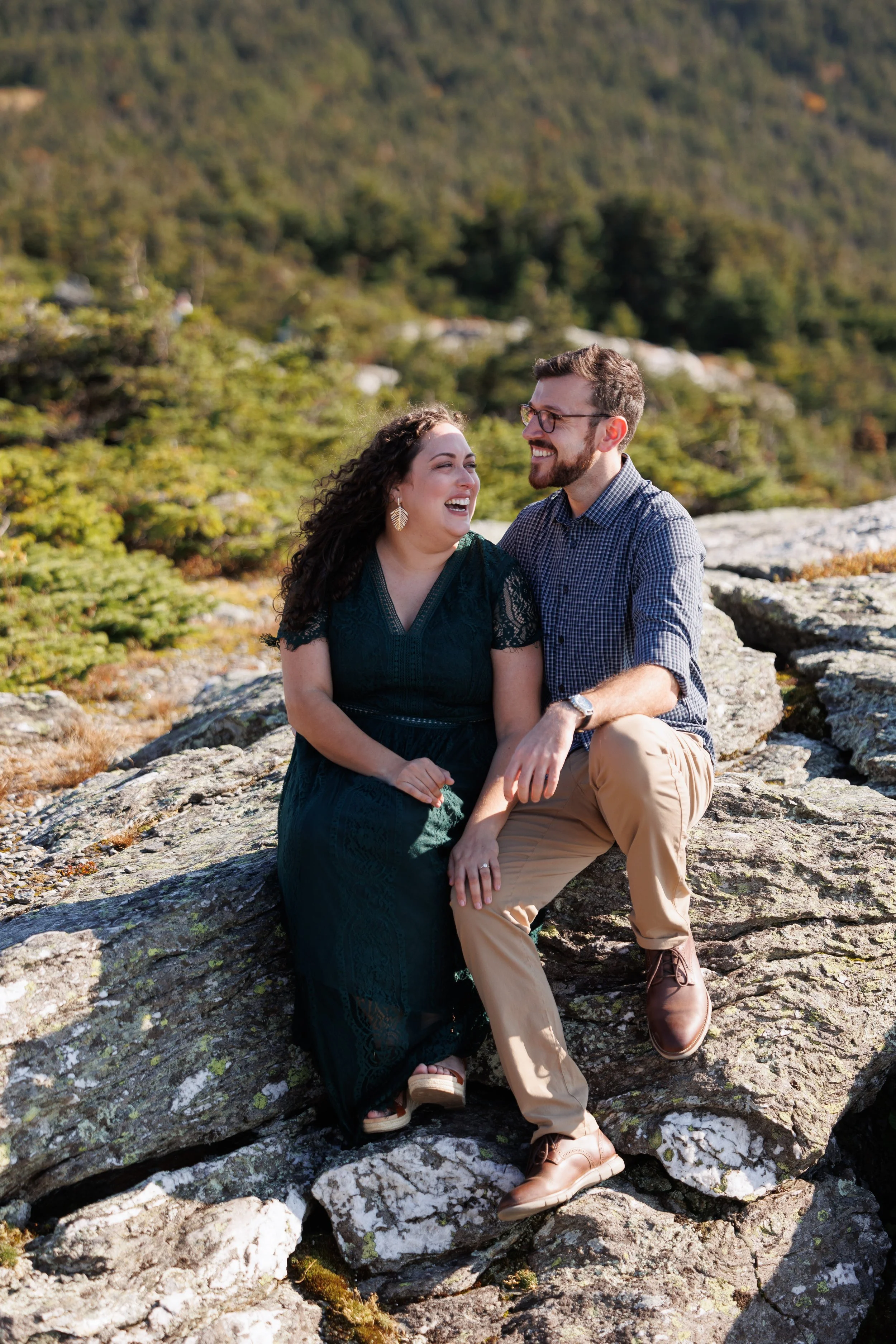 A smiling couple sitting on rocks outdoors with a forested hillside background, enjoying a sunny day.