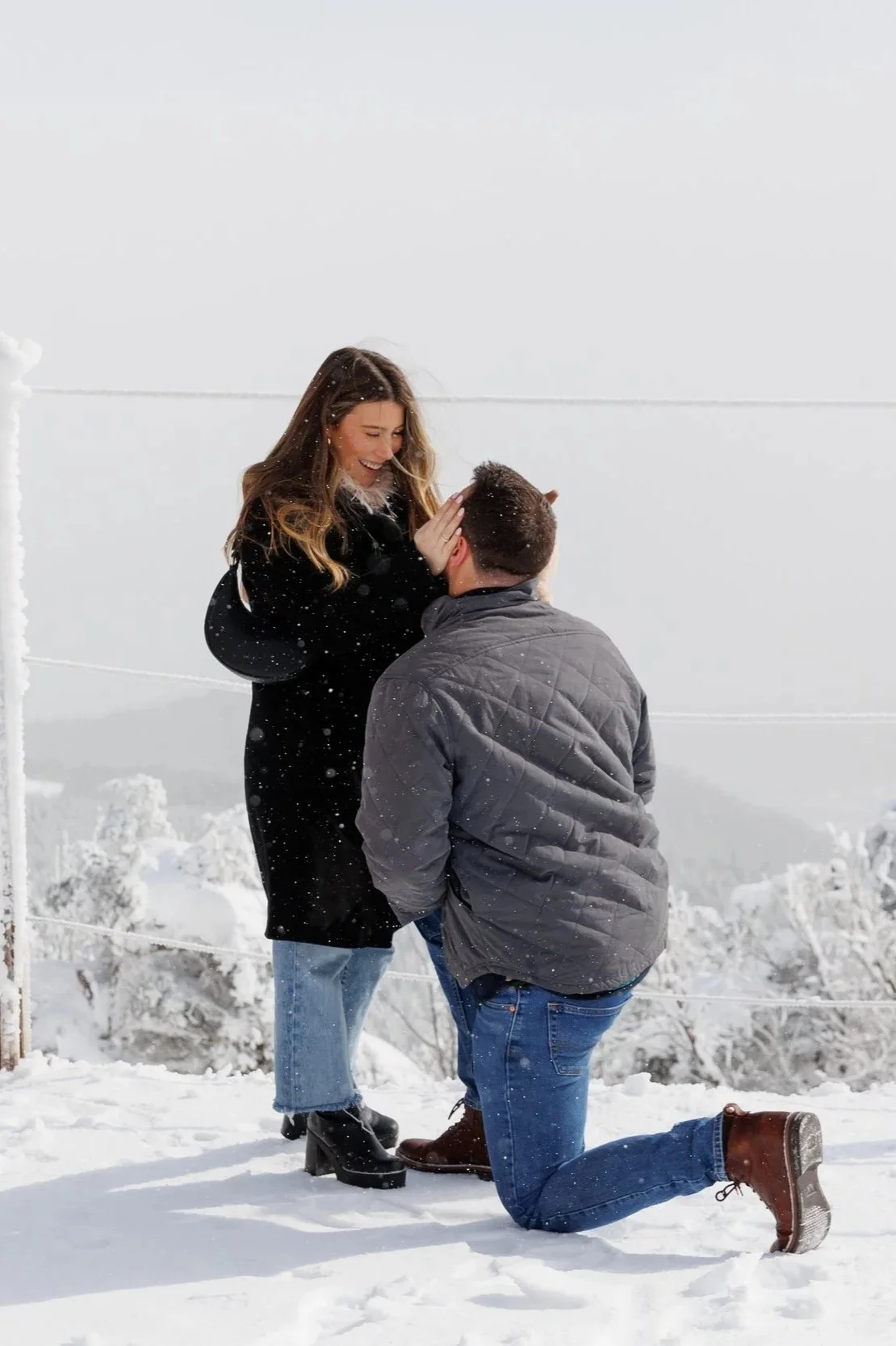 A woman is proposing to a man on a snowy outdoor landscape, with her holding his face and smiling as he kneels on one knee.