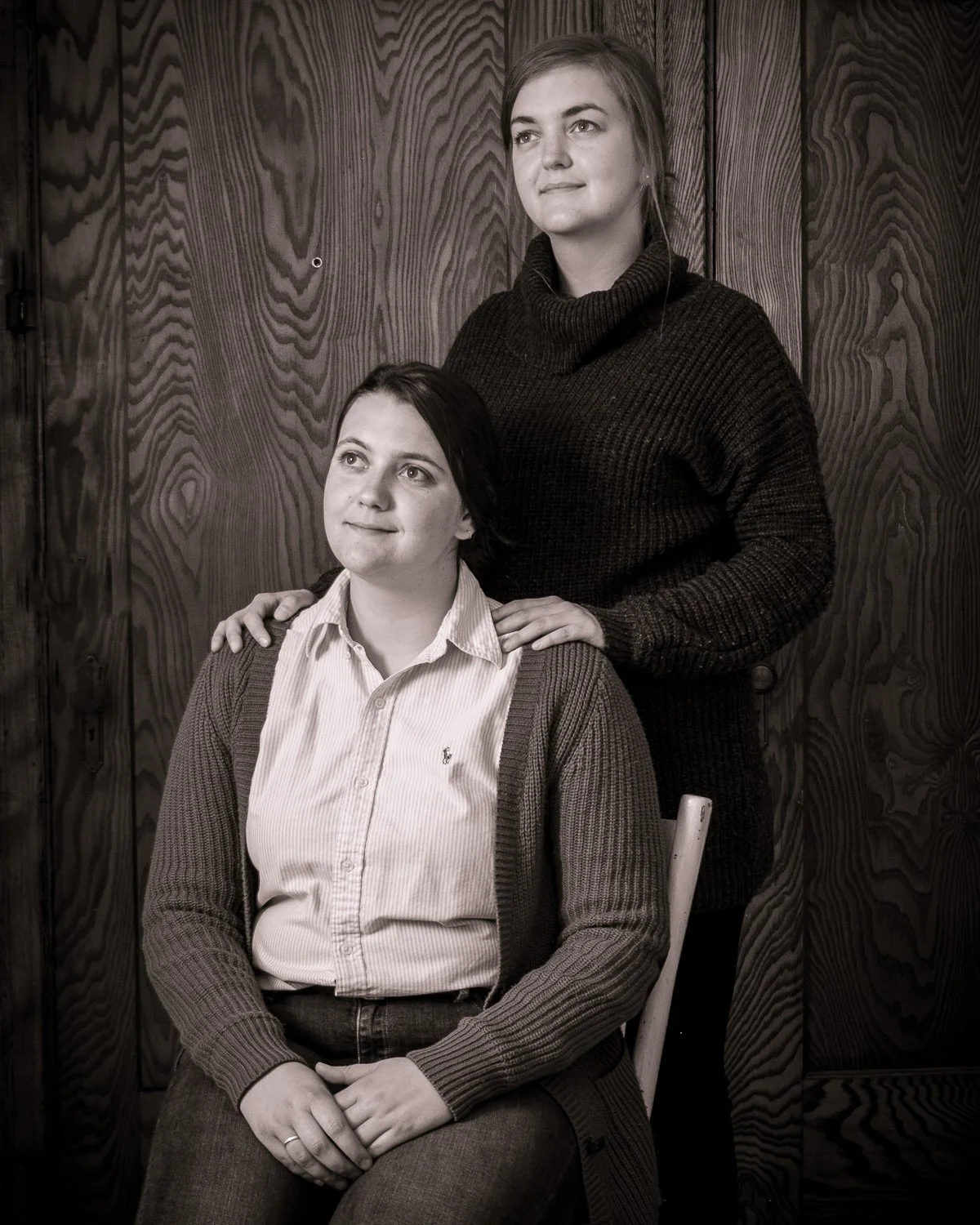 Deux femmes, l'une assise et l'autre debout derrière, posant devant un mur en bois, en noir et blanc.