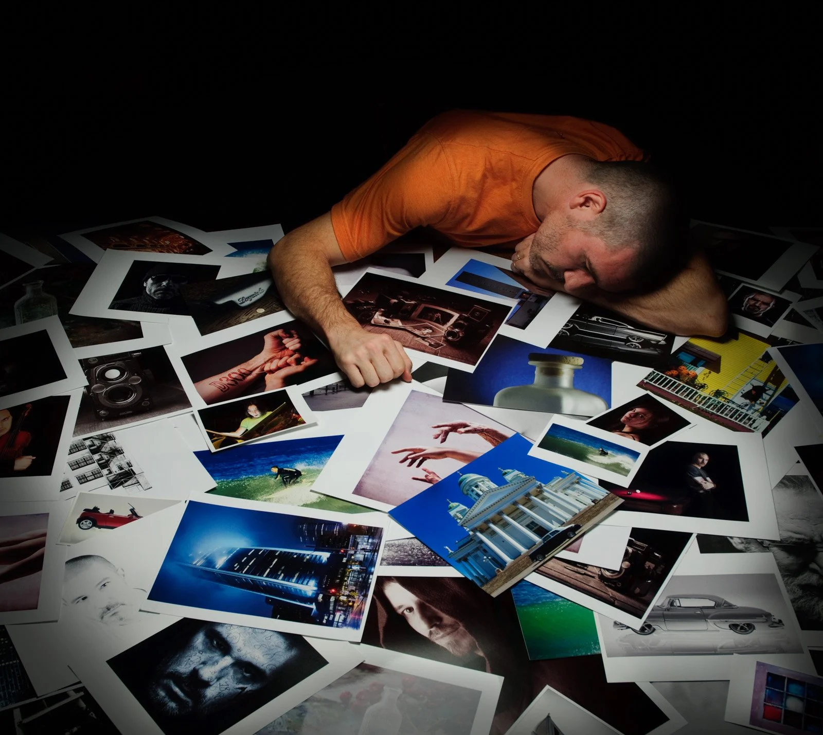 Un homme allongé sur une table avec une tête reposant sur son bras, entouré de nombreuses photographies dispersées, représentant différentes scènes, personnes, et objets.