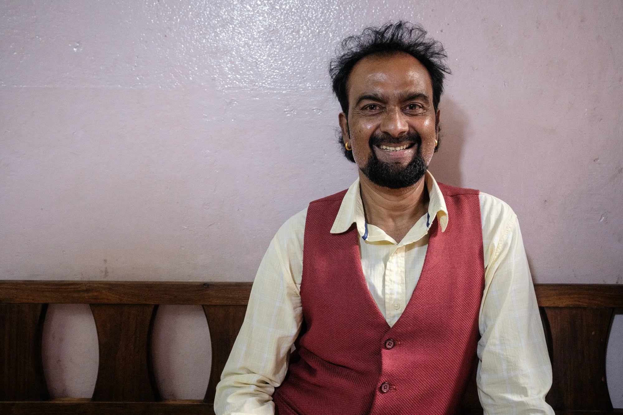 Un homme souriant, avec des cheveux noirs et une barbe, portant une veste rose et une chemise beige, assis devant un mur blanc