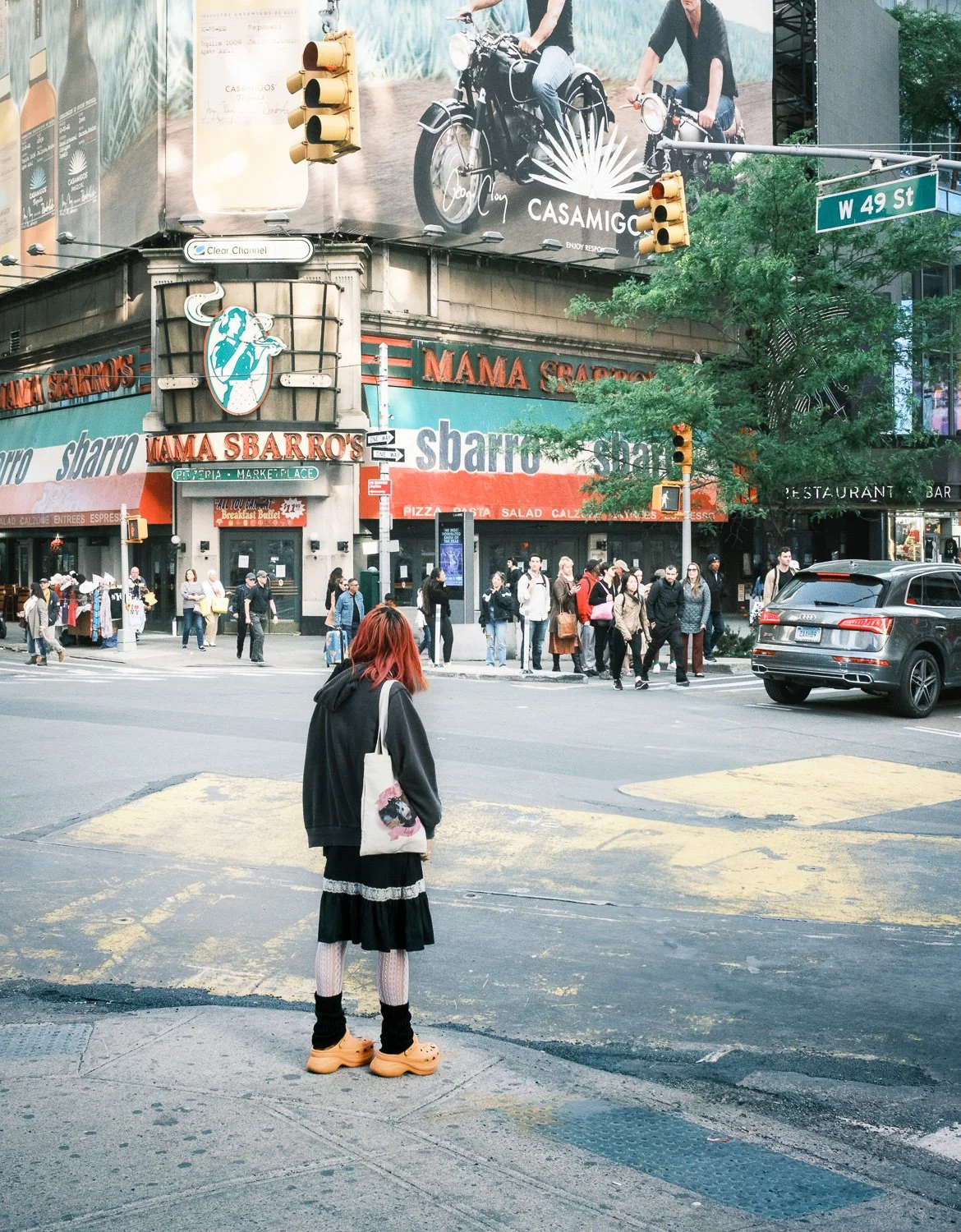 Une scène urbaine à Times Square avec plusieurs piétons qui traversent la rue, un restaurant nommé Mama Sbarro, des panneaux publicitaires en arrière-plan, et un feu de circulation. Au premier plan, une personne avec des cheveux rouges, portant un sw