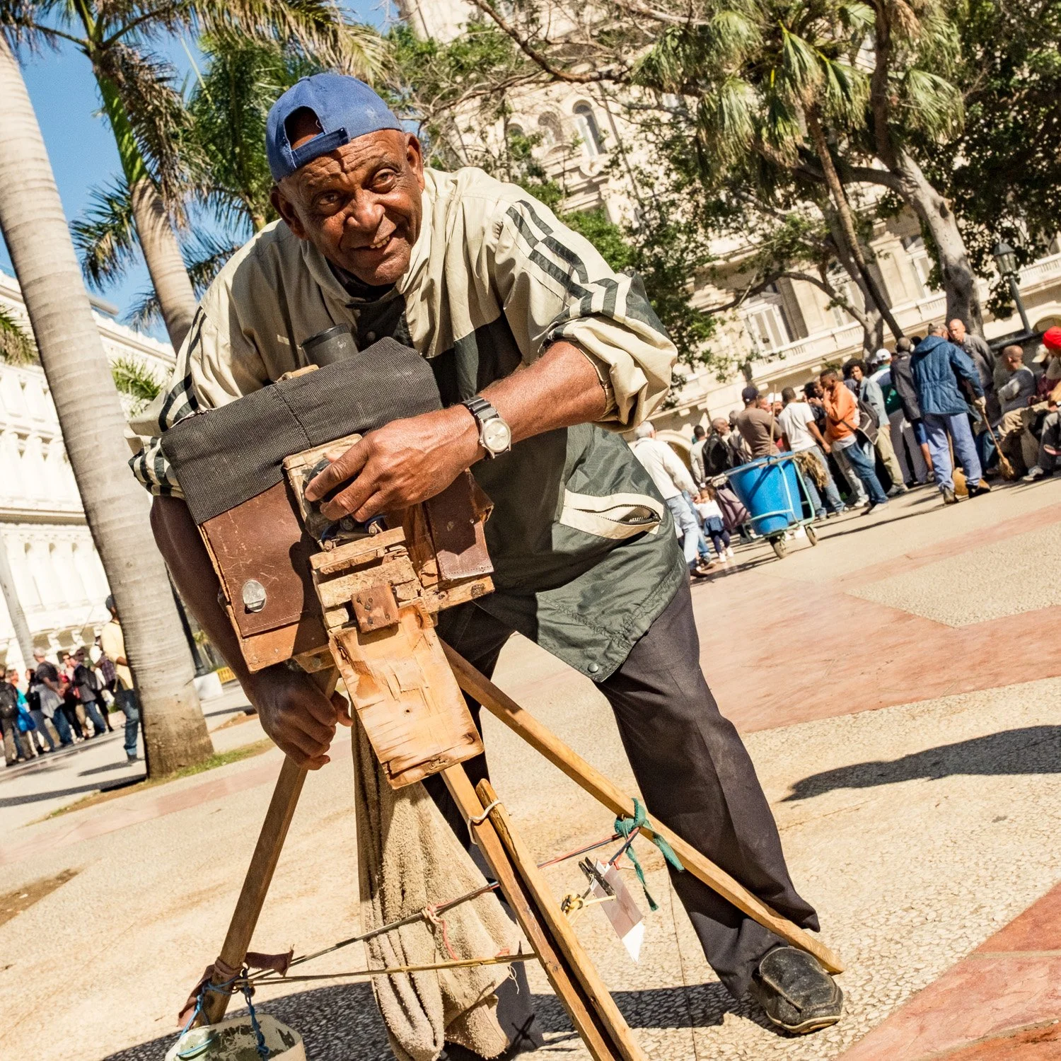 Un homme âgé souriant en train de réaliser une photographie de rue avec un équipement vintage, dans un lieu public en plein air, entouré de personnes et d'arbres, par une journée ensoleillée.