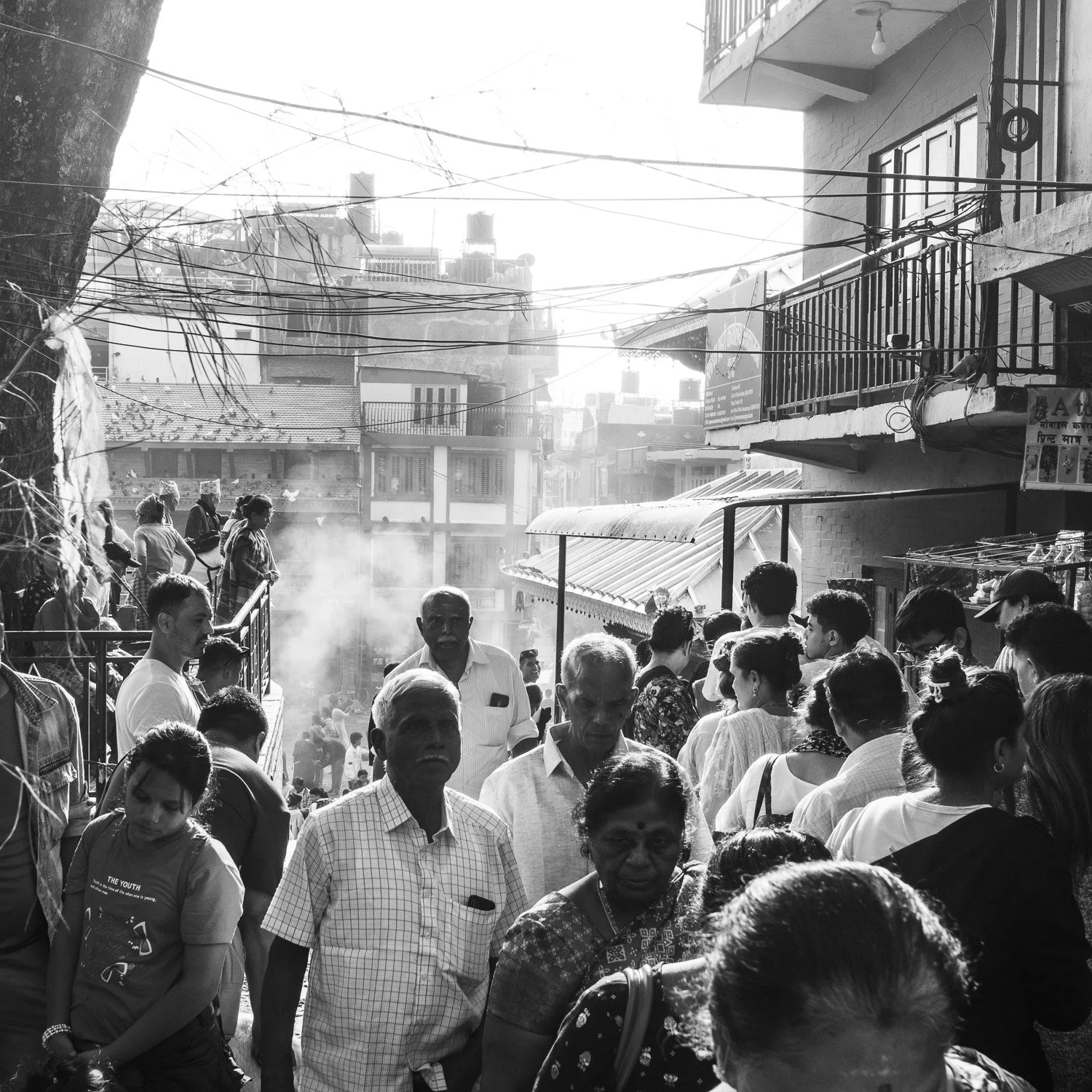 Une foule de personnes dans une rue urbaine en Inde, avec des bâtiments en arrière-plan, certains fumant de la vapeur ou de la fumée. La majorité des personnes portent des vêtements traditionnels ou modernes et semblent attendre ou observer quelque c