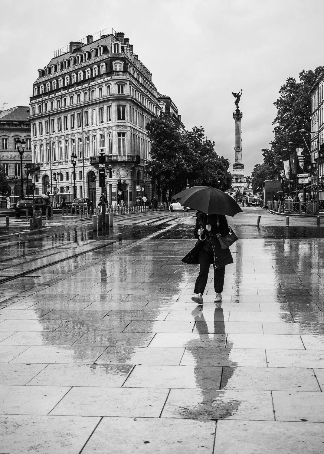 Une femme marche sous un parapluie noir sur un trottoir mouillé à Paris, avec des bâtiments historiques et une statue en arrière-plan, par temps pluvieux.