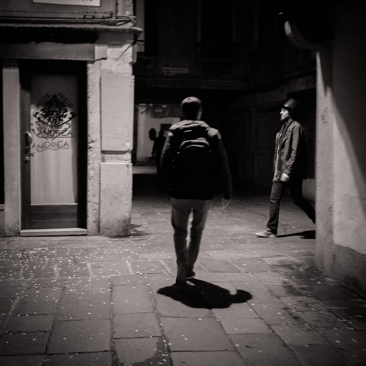 Deux hommes marchent dans une rue sombre la nuit, l'un porte un chapeau haut de forme et un manteau, l'autre porte un sac à dos.