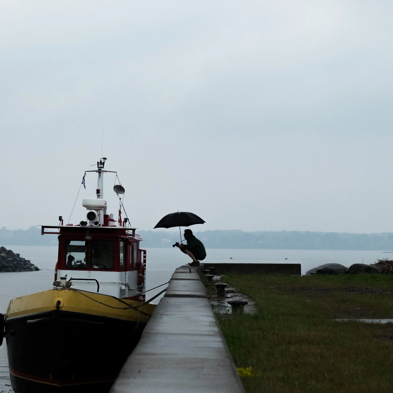 Une personne assise sur un quai en pierre, sous un parapluie noir, en train de prendre une photo, avec un bateau rouge et blanc amarré près du quai, et une vue sur un lac ou une mer dans un paysage brumeux ou pluvieux.