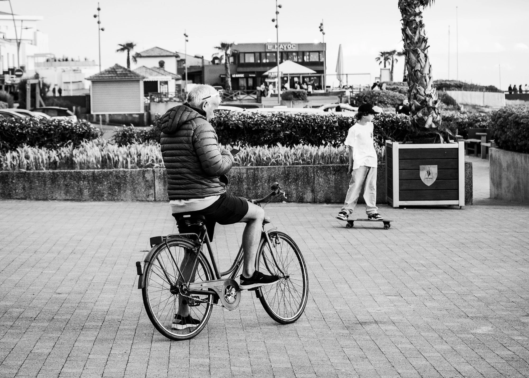 Une personne à vélo portant une veste courte et un pantalon court passe devant un garçon skateboarder sur un trottoir face à une zone piétonne avec des palmiers et un bâtiment en arrière-plan.