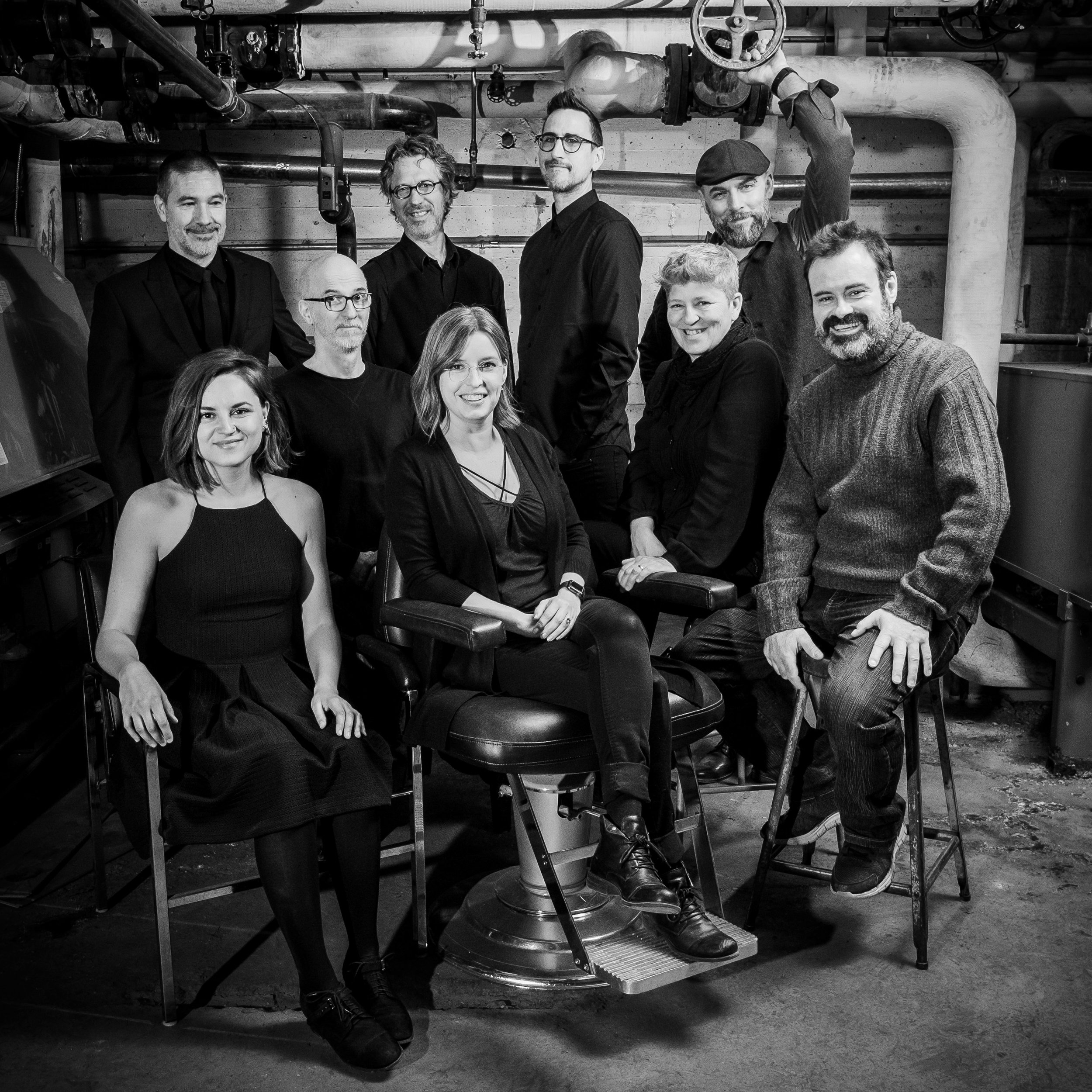 Groupe de huit personnes posant pour une photo dans un environnement industriel, certains assis sur des chaises et d'autres debout, tous souriants, en noir et blanc.