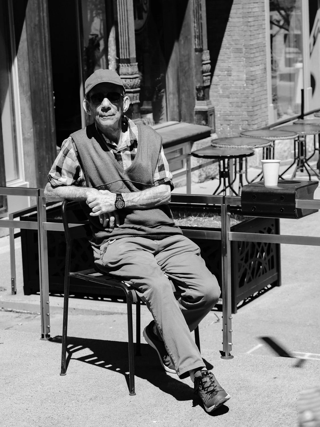Homme âgé assis sur une chaise dehors, portant des lunettes de soleil, une casquette, une chemise à carreaux, un pull sans manches et un pantalon, avec une tasse en papier sur une table à côté de lui.